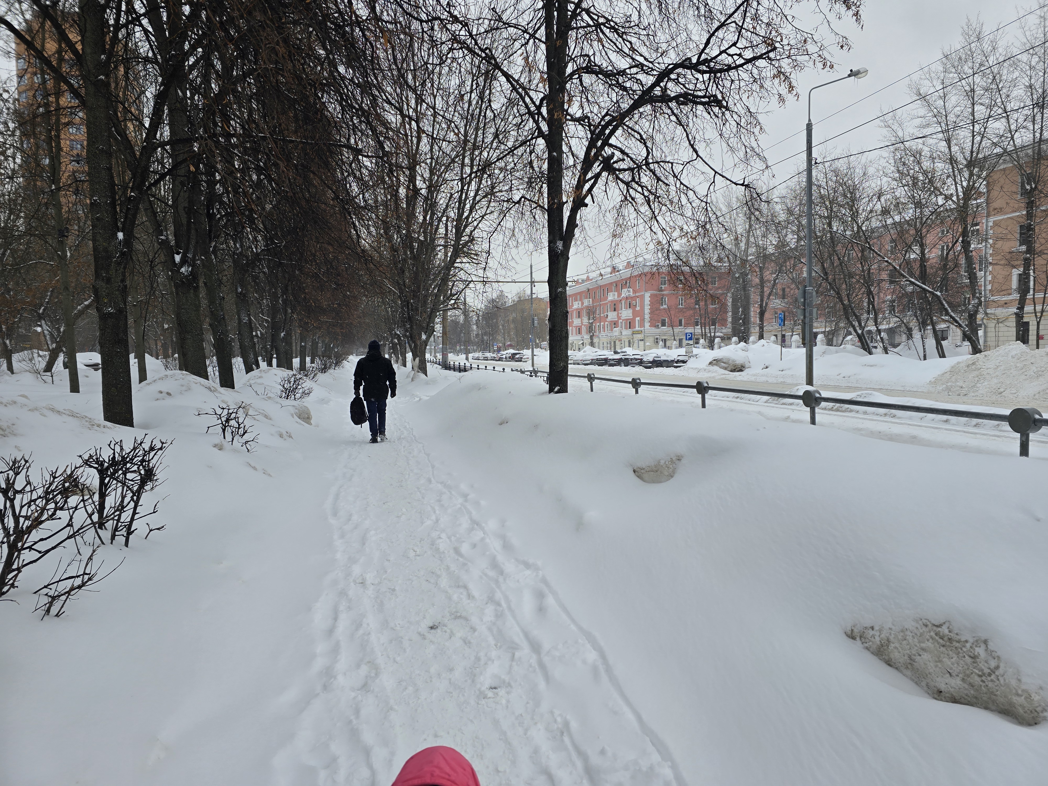 Участок городской дороги с пешеходной дорожкой, отделенной от проезжей части металлическим ограждением. Дорога и тротуар покрыты снегом, переходов в кадре не видно.