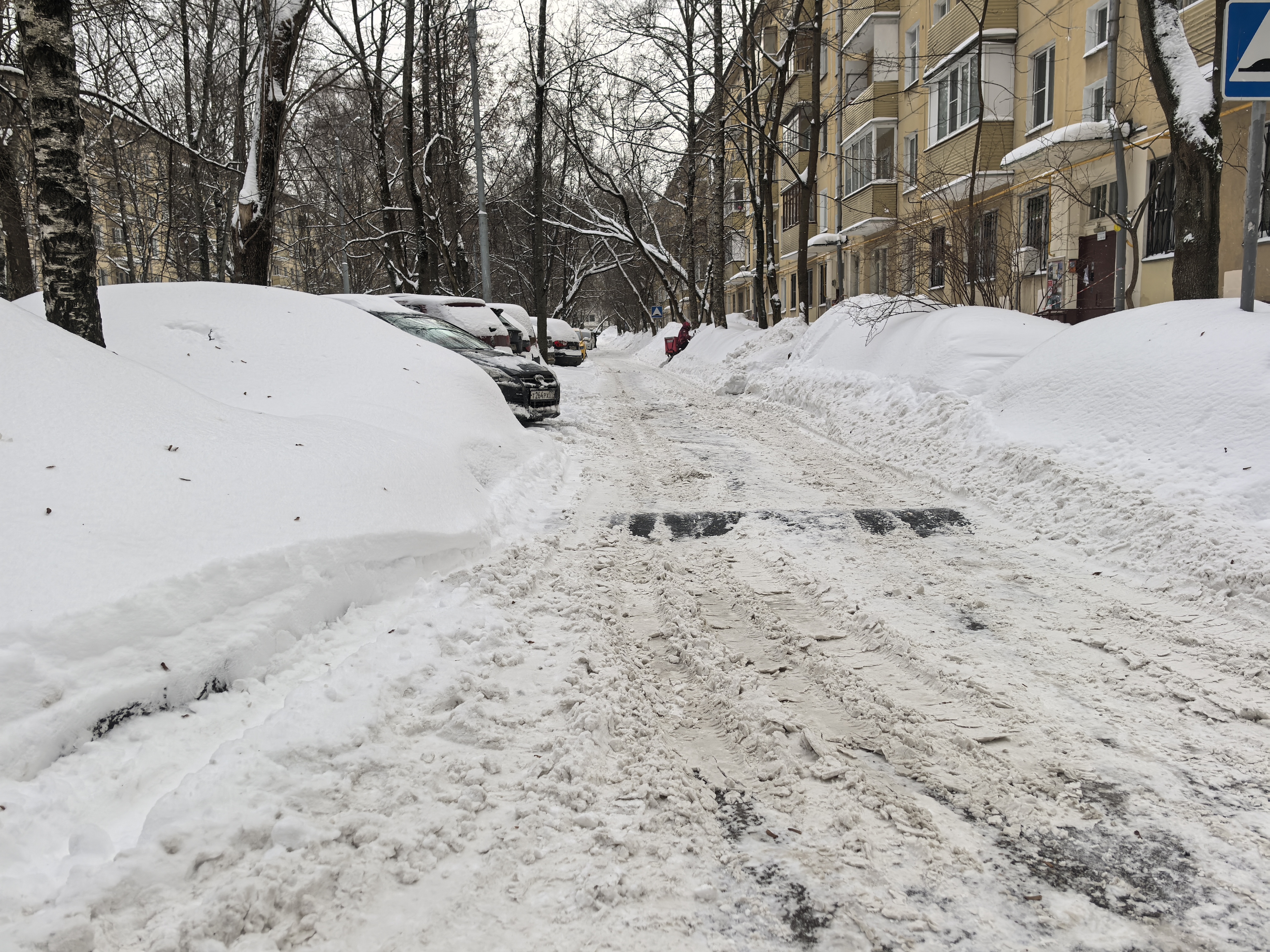 Дворовая территория, заснеженная дорога между жилыми домами и деревьями. Проезжая часть очищена частично, покрыта снегом и наледью. Видны припаркованные автомобили, частично засыпанные снегом.