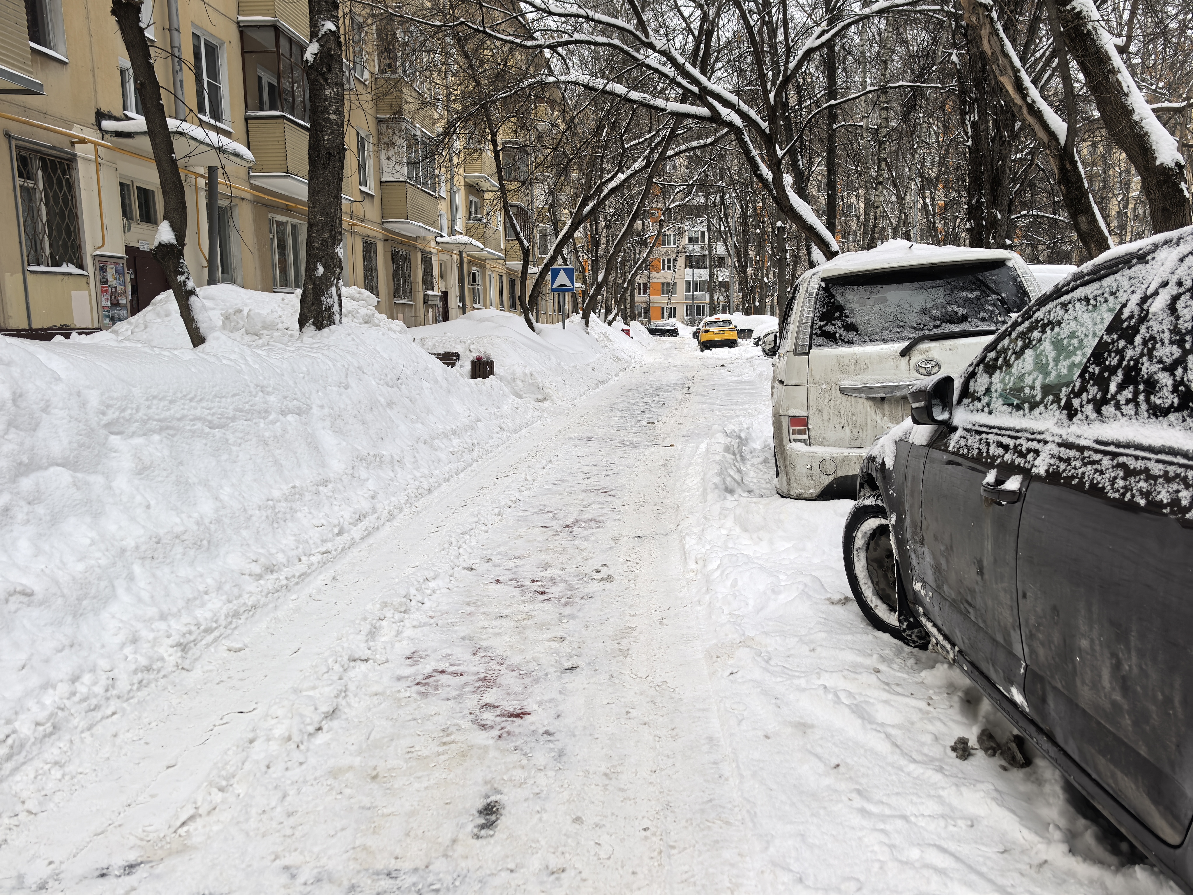 Дворовая территория с припаркованными автомобилями, покрытая снегом. Видны жилые дома и деревья. Проезжая часть узкая, очищена от снега частично.