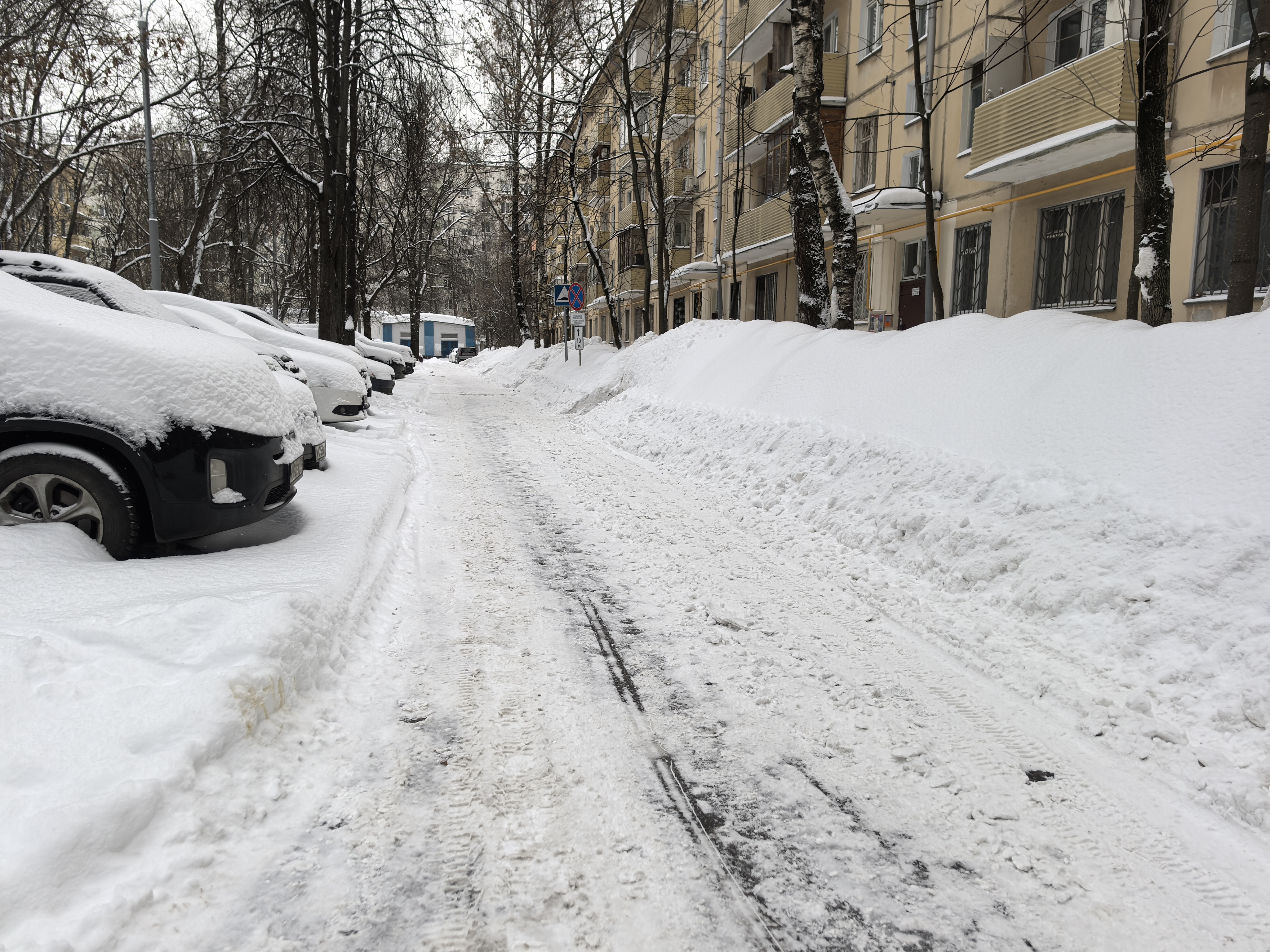 Дворовая территория с припаркованными автомобилями, покрытая снегом. Проезжая часть узкая, тротуары полностью засыпаны снегом.