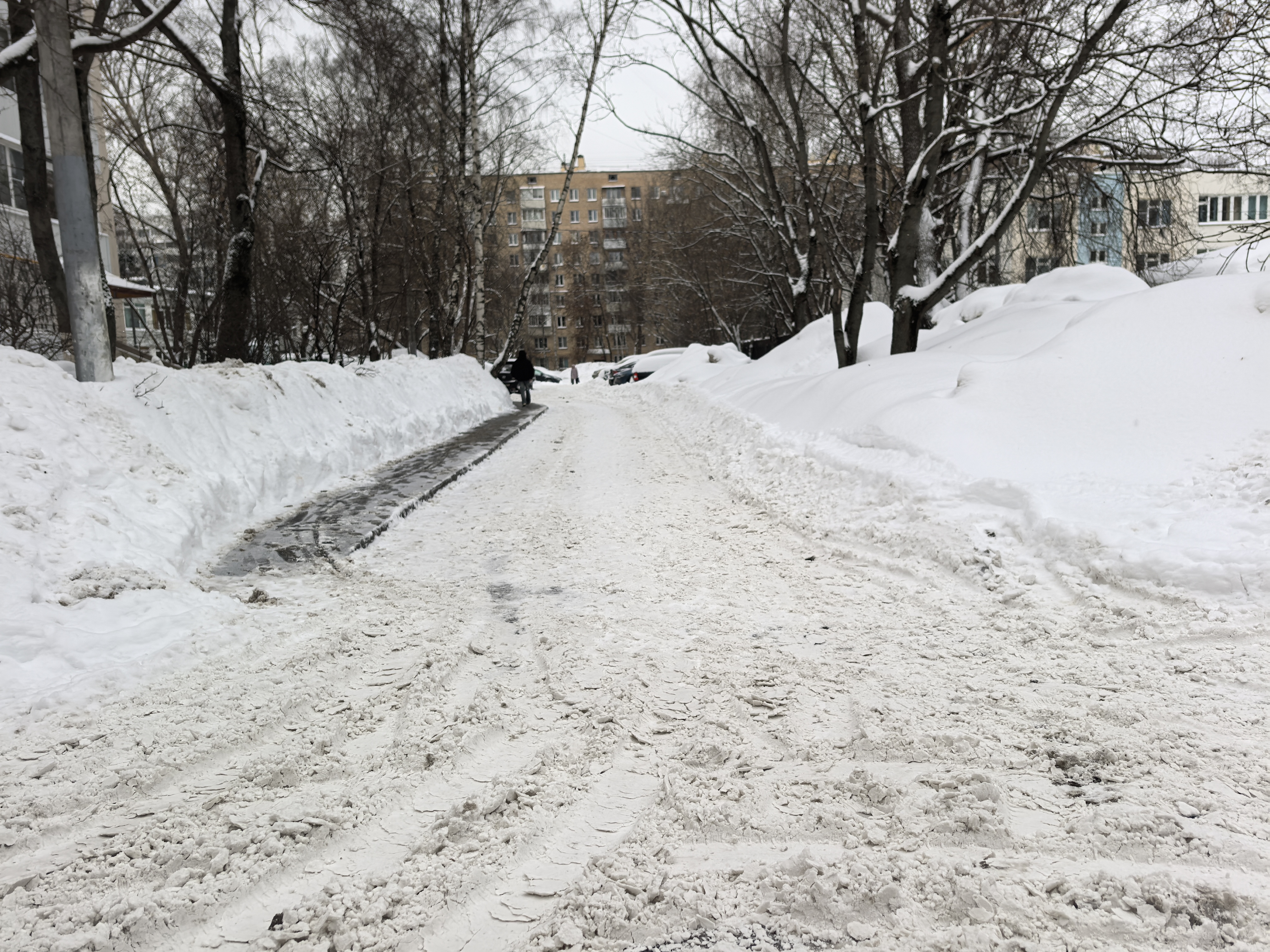Дворовая территория с заснеженной дорогой, окруженная жилыми домами и деревьями. Тротуарная дорожка слева узкая и скользкая, основная проезжая часть покрыта снегом и следами шин.