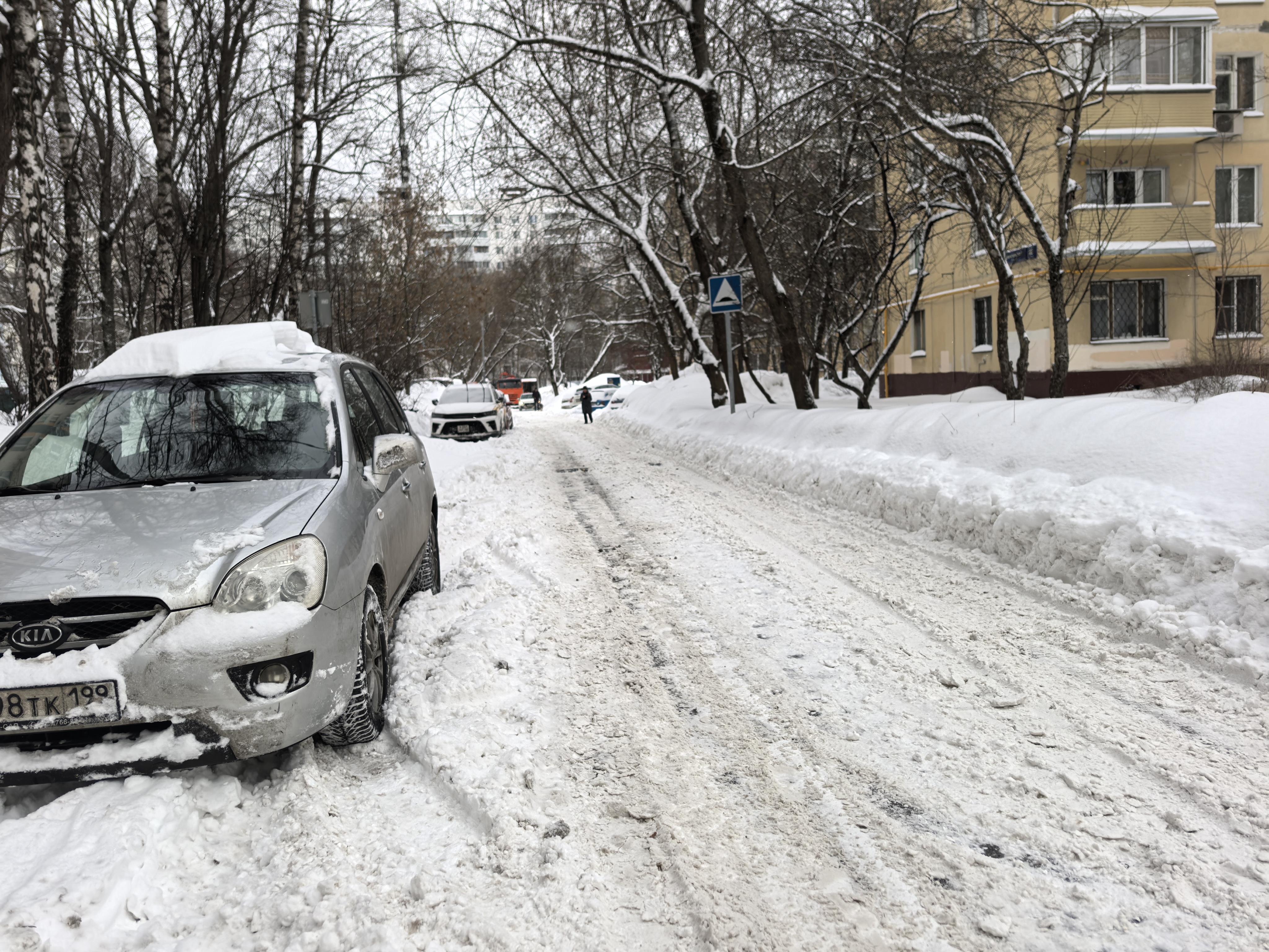 Участок городской улицы с проезжей частью, покрытой снегом и колеей. Пешеходный переход отсутствует, но виден знак, указывающий на его наличие впереди. Тротуары полностью засыпаны снегом.