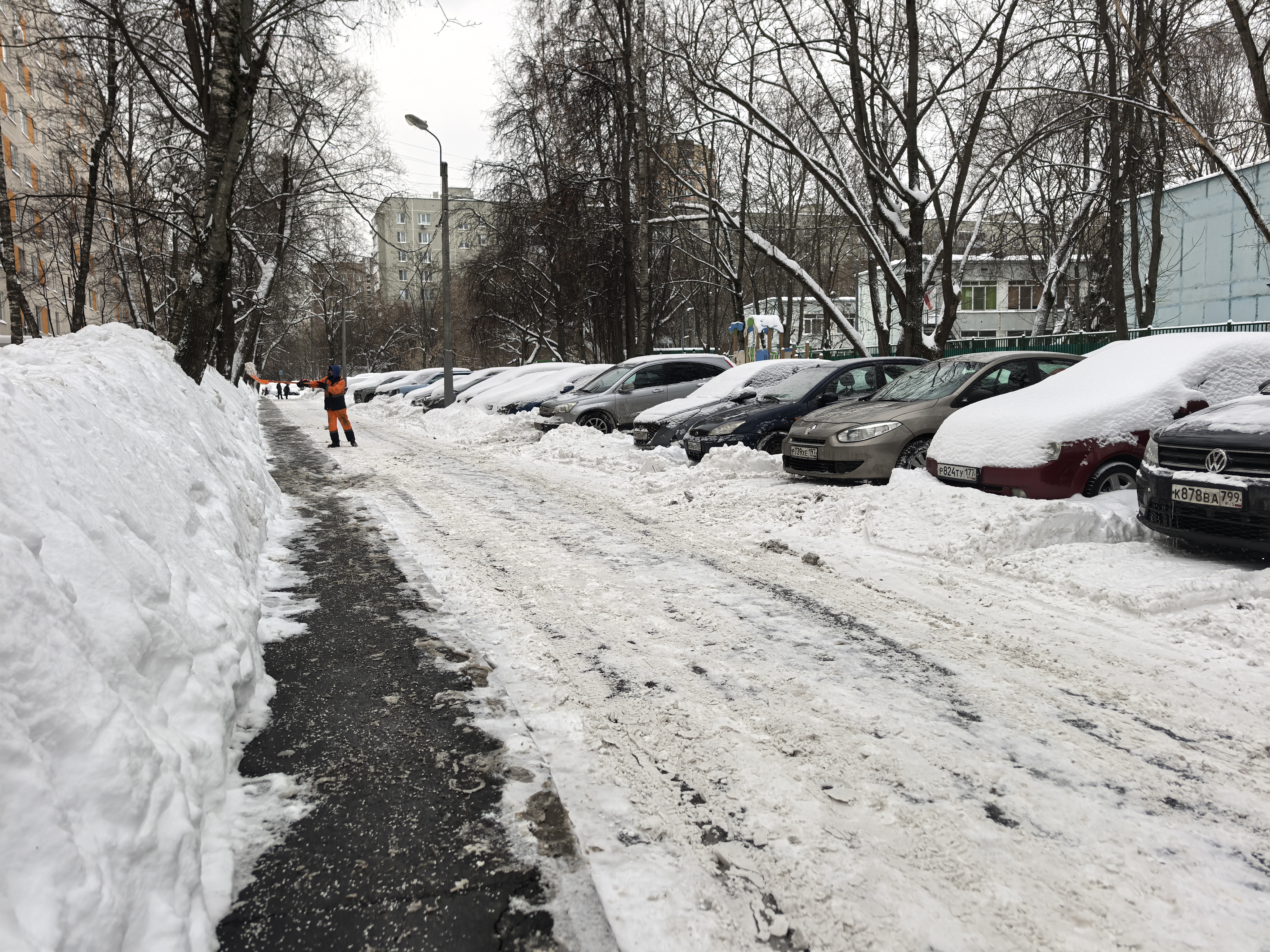 Дворовая территория (парковка/проезд между домами), покрытая снегом, с припаркованными автомобилями и узким пешеходным тротуаром.
