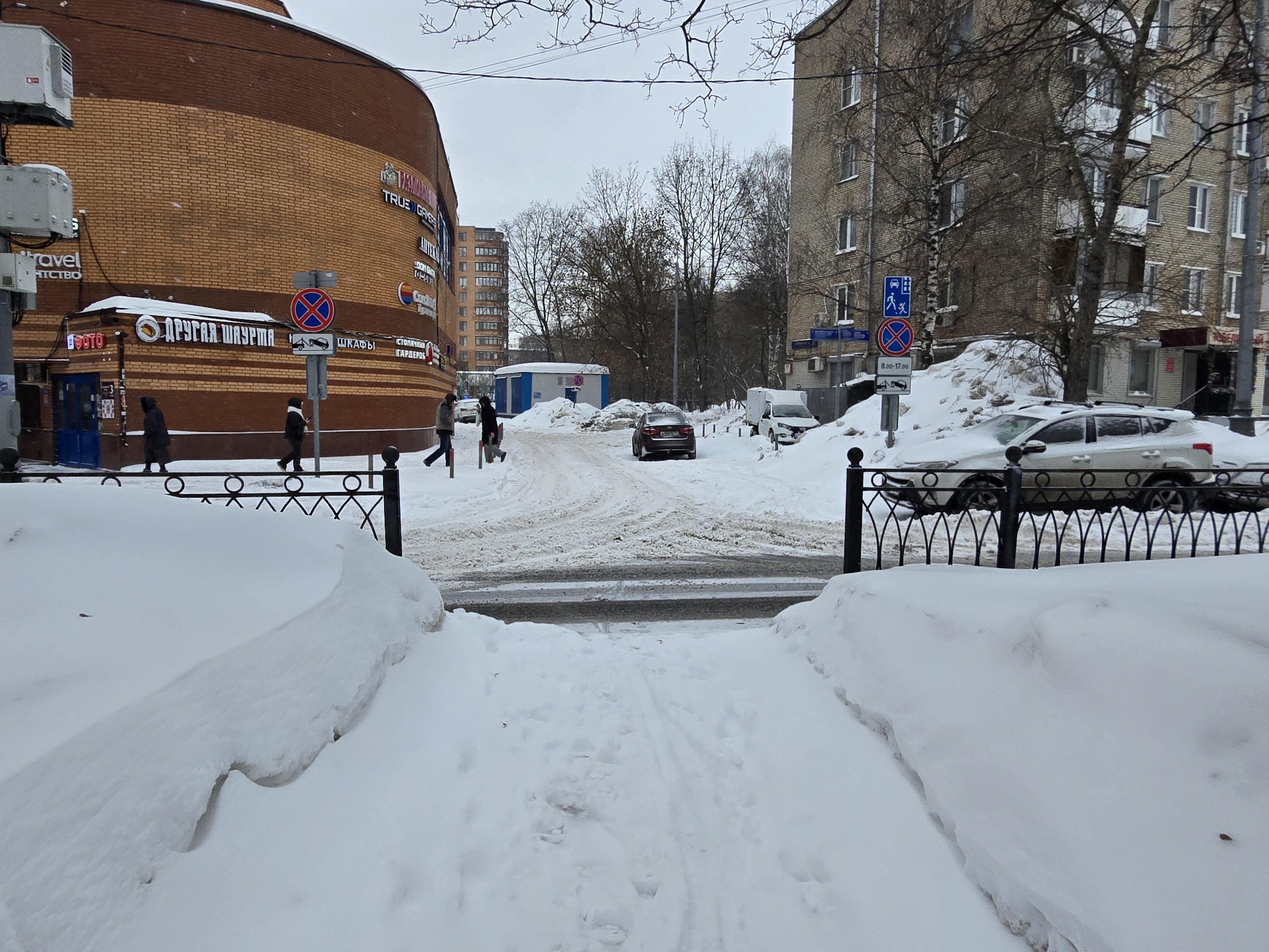 Участок проезжей части без пешеходного перехода, покрытый снегом, с припаркованными автомобилями и пешеходами, идущими по обочине.