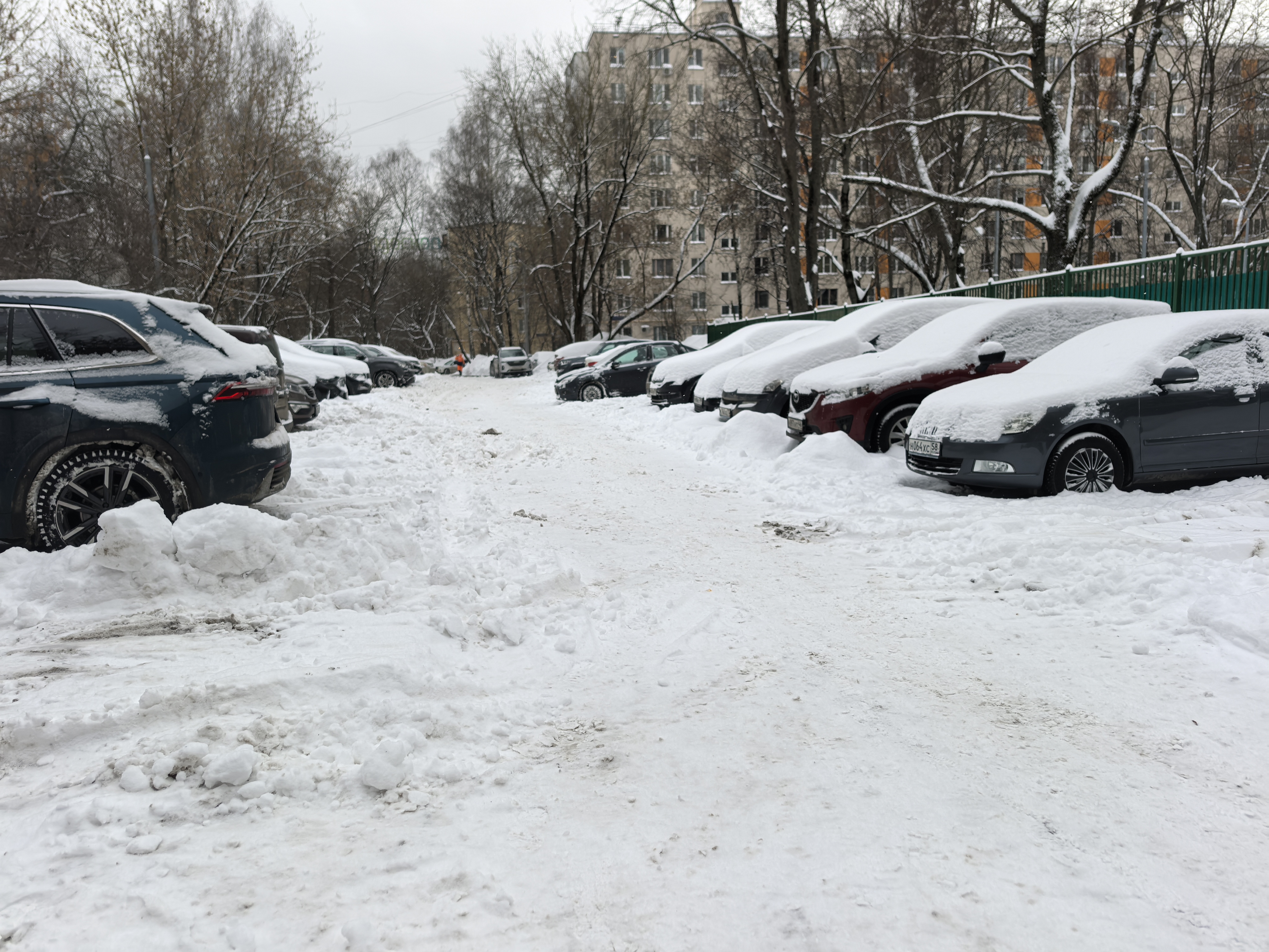 Дворовая территория, используемая как парковка, покрытая снегом. Видны припаркованные автомобили с обеих сторон проезжей части, жилые дома на заднем плане.