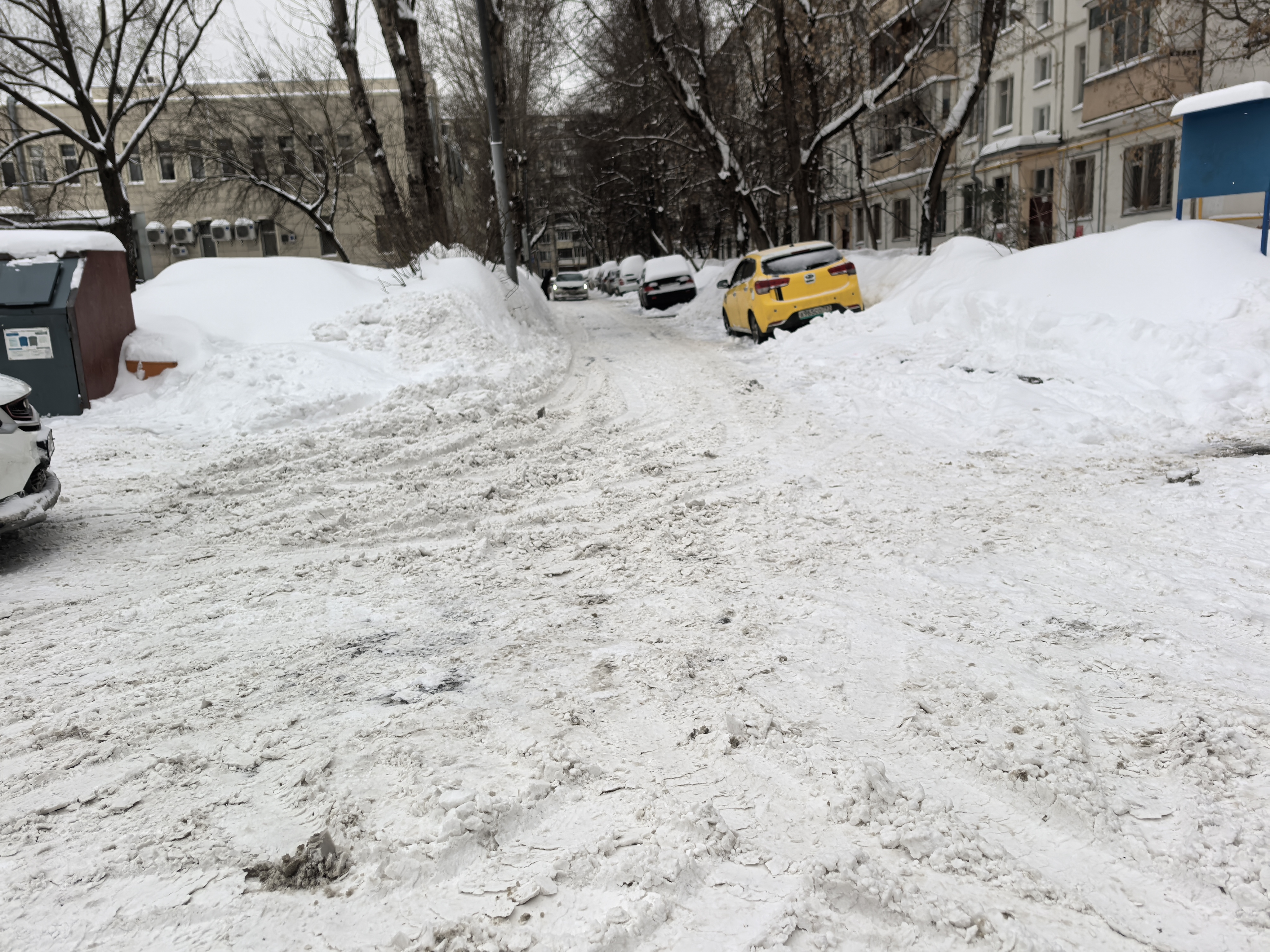 Дворовая территория с припаркованными автомобилями и заснеженной дорогой, тротуары отсутствуют или полностью засыпаны снегом.