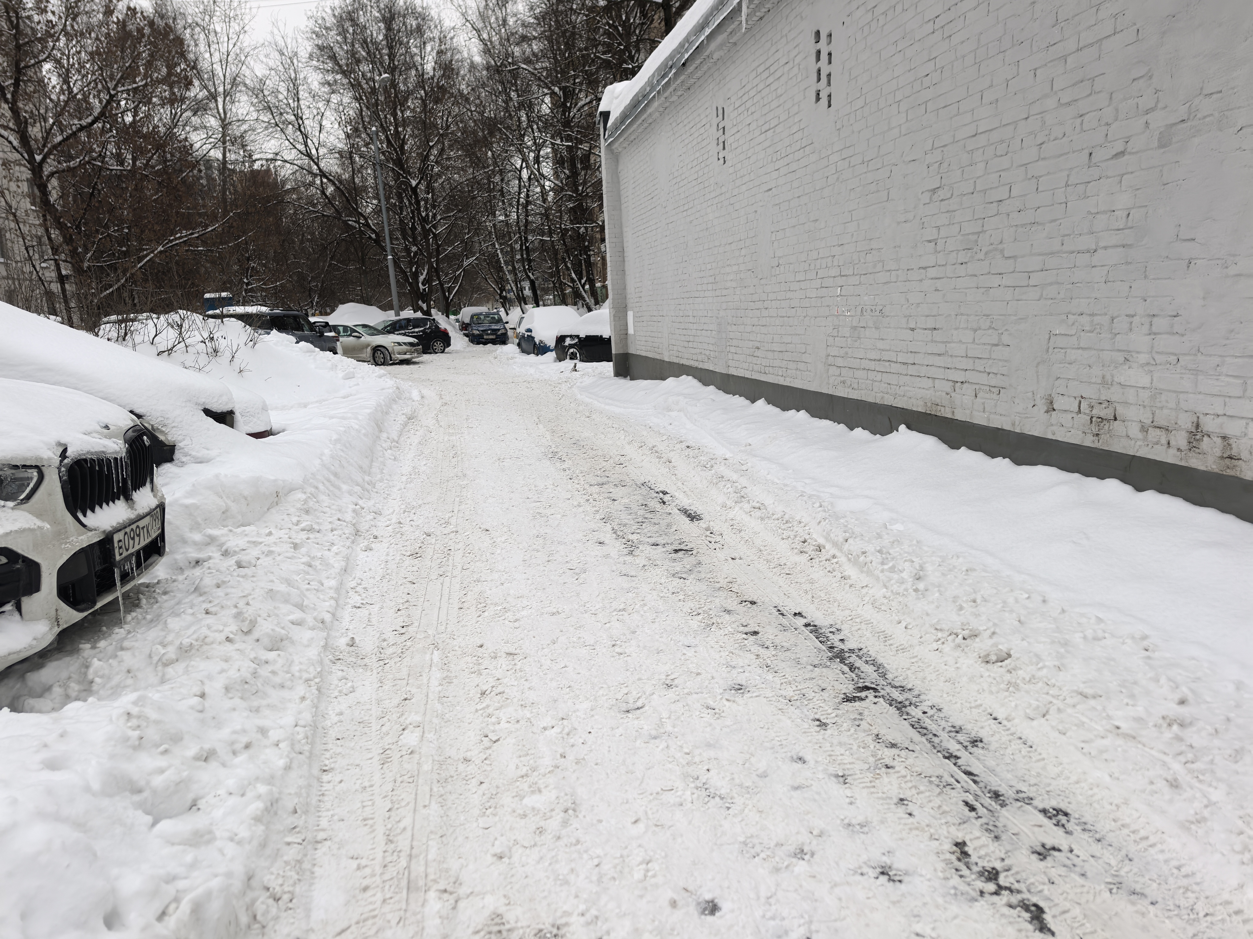 Дворовая территория, заснеженная дорога с припаркованными автомобилями по обеим сторонам, видна белая кирпичная стена справа и деревья слева.