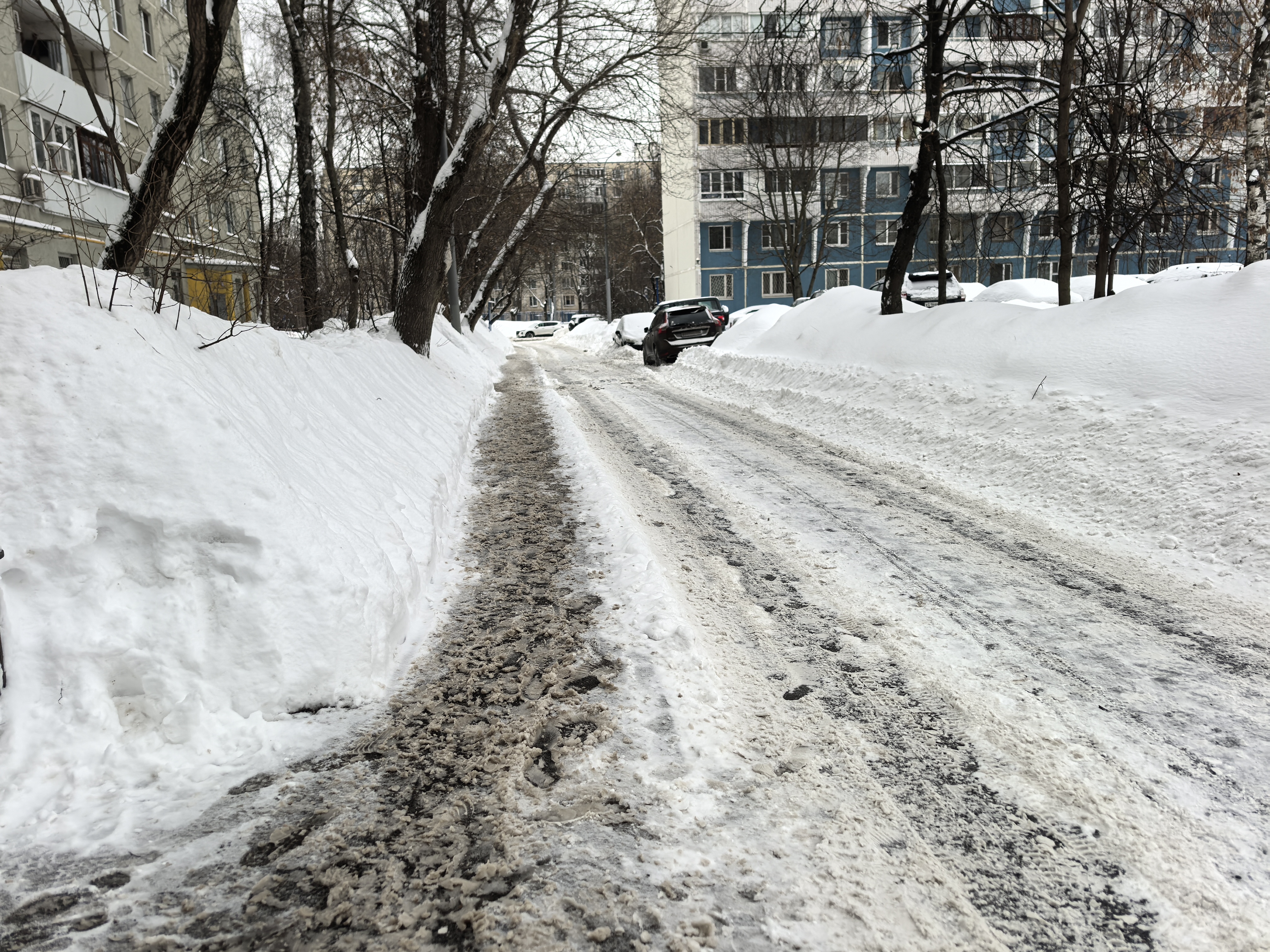 Дворовая территория, зимний период, дорога покрыта снегом и наледью, припаркованные автомобили вдоль проезжей части, тротуары полностью засыпаны снегом.