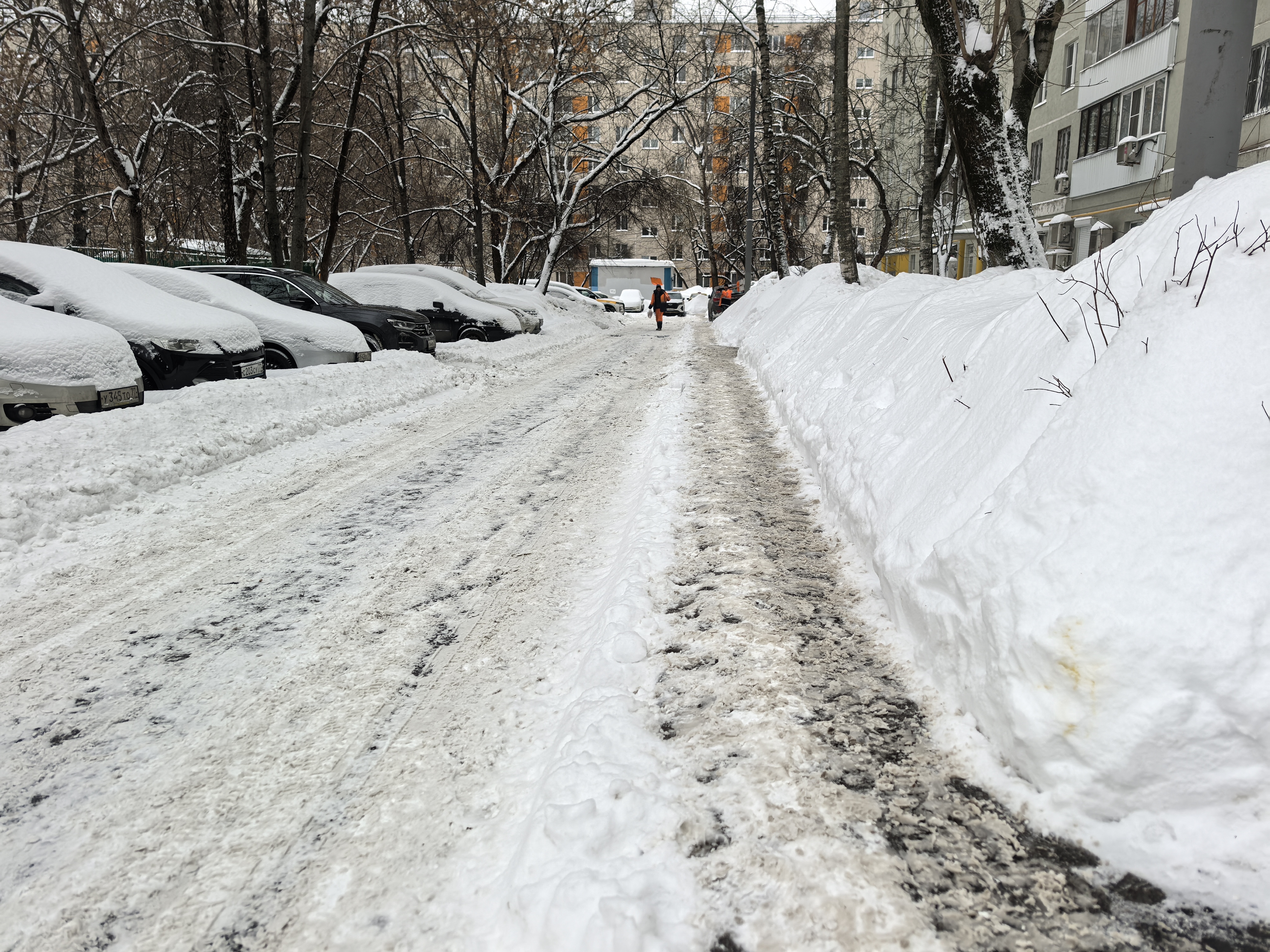 Дворовая территория с припаркованными автомобилями, покрытая снегом и ледяной коркой. Видны жилые дома и деревья.
