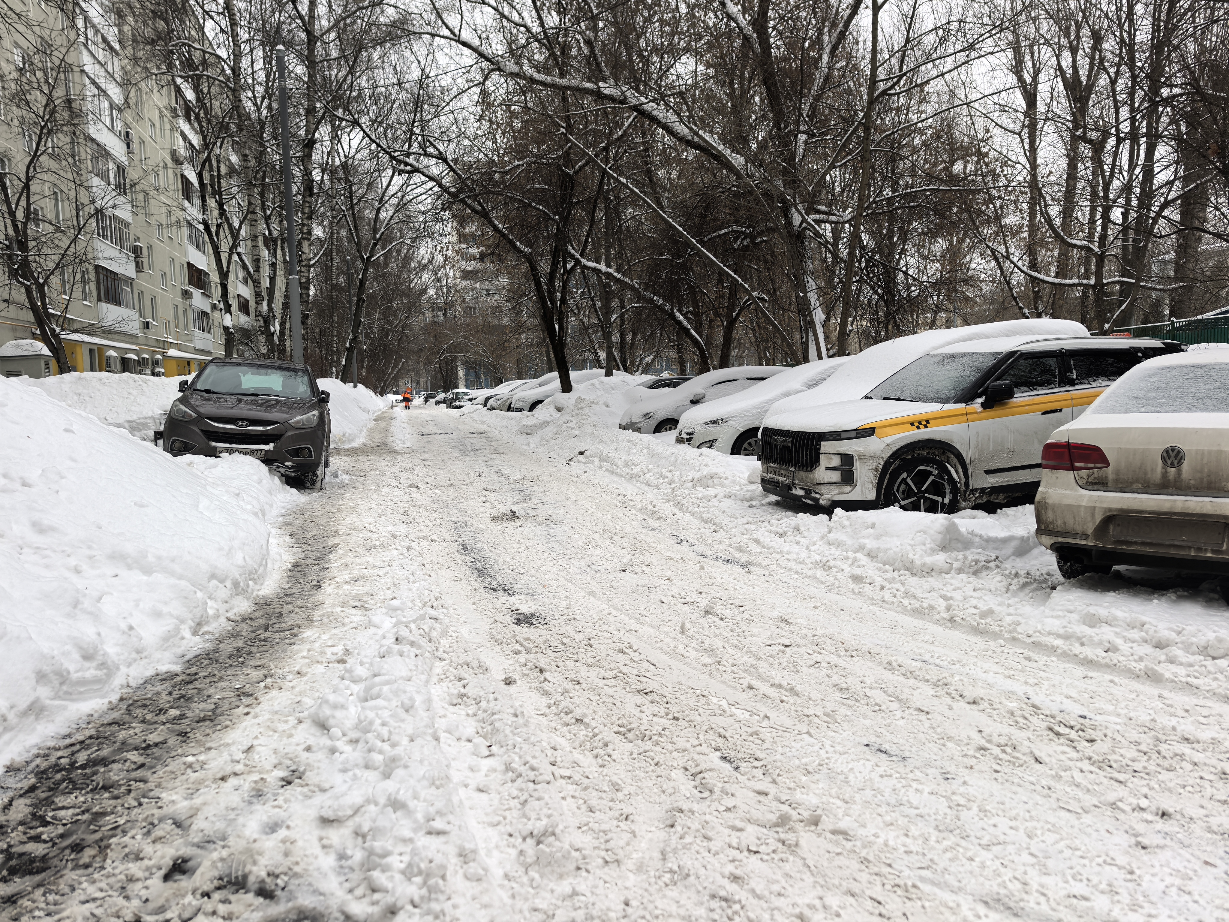 Дворовая территория, узкая проезжая часть, ограниченная припаркованными автомобилями и сугробами, покрытая снегом и наледью. Видны жилые дома и деревья.