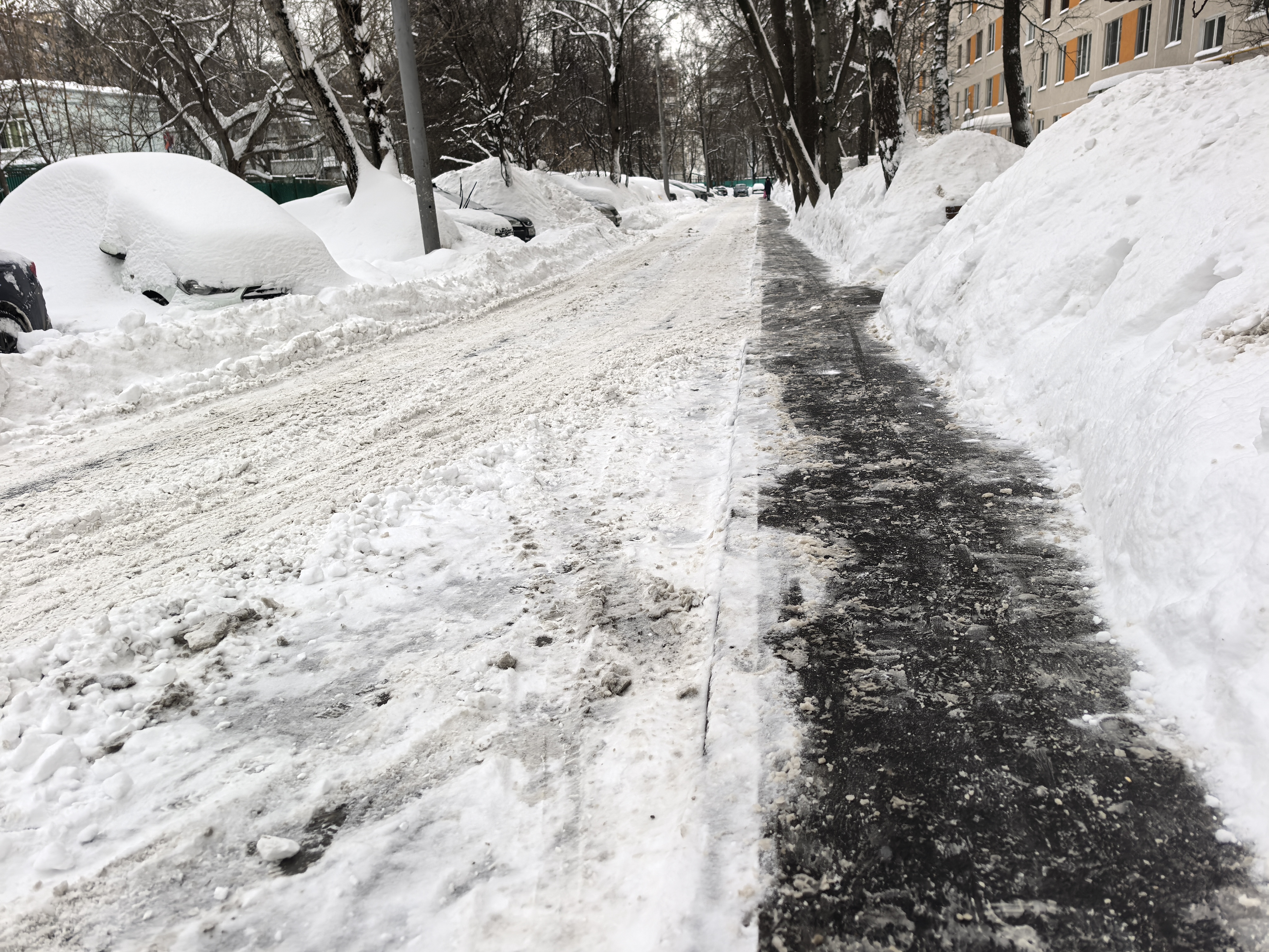 Участок дороги без пешеходного перехода, с узким тротуаром, покрытым льдом и снегом, и припаркованными автомобилями, частично скрытыми снегом.