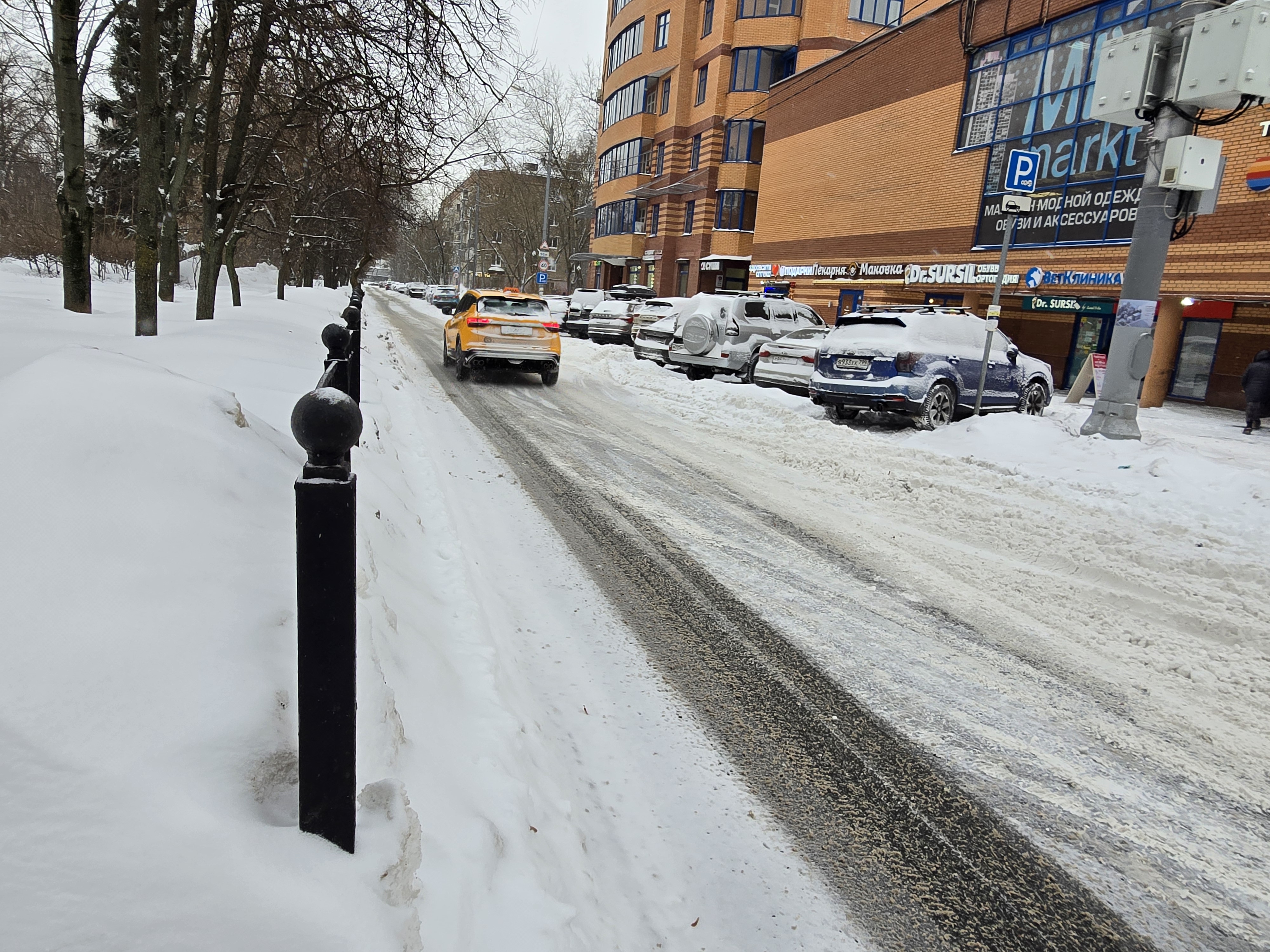 Участок городской улицы с проезжей частью, покрытой снегом и льдом. Справа — припаркованные автомобили у здания, слева — тротуар, полностью засыпанный снегом. Пешеходный переход на видимом участке отсутствует.