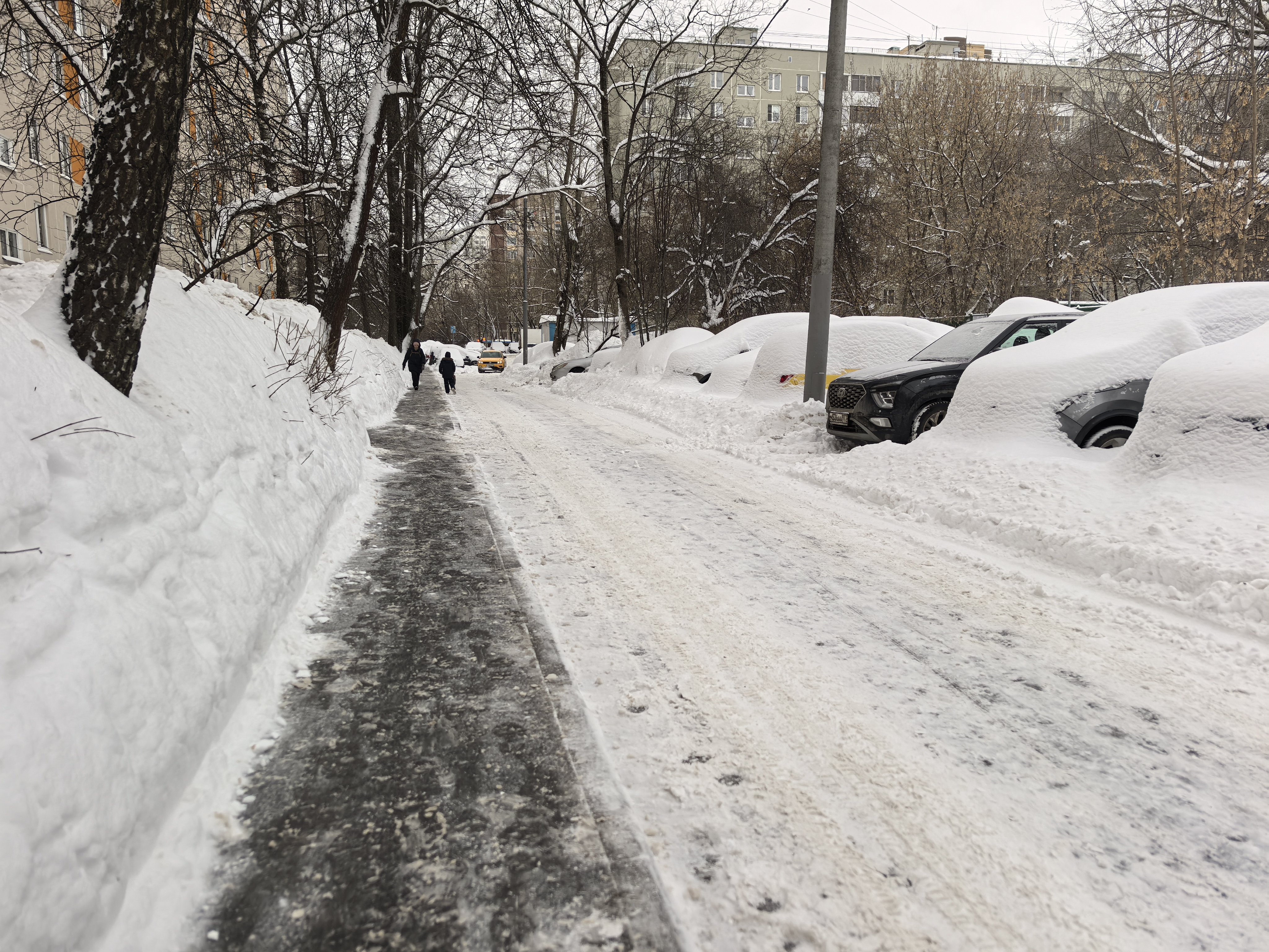 Участок городской улицы зимой. Видна проезжая часть, покрытая снегом и льдом, и узкий тротуар, очищенный от снега. Справа припаркованы автомобили, полностью засыпанные снегом. На тротуаре идут пешеходы. Пешеходный переход на видимом участке отсутствует.