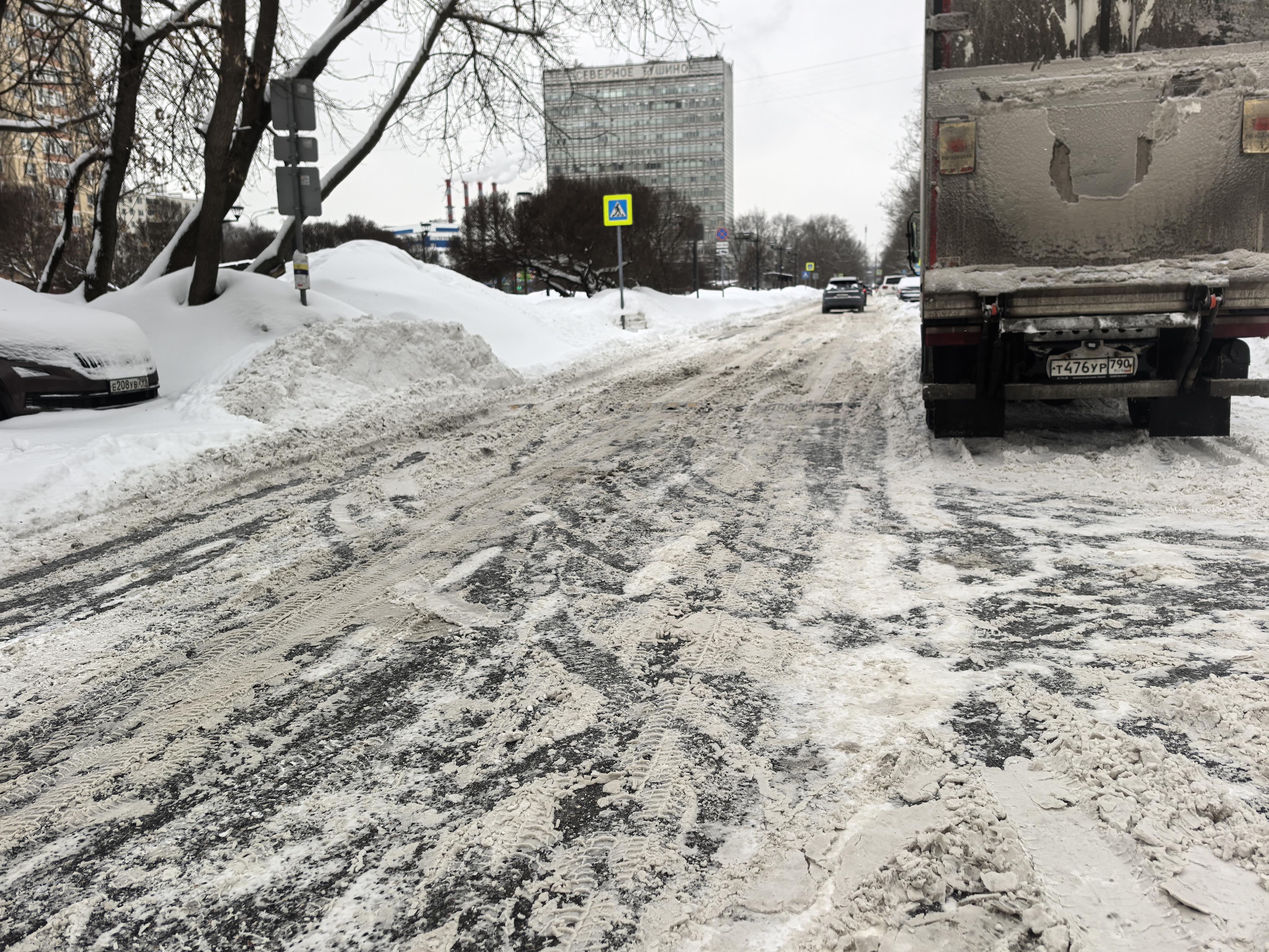Участок проезжей части без пешеходного перехода (зебры), покрытый снегом и слякотью. Видны припаркованные автомобили и движущийся грузовик.