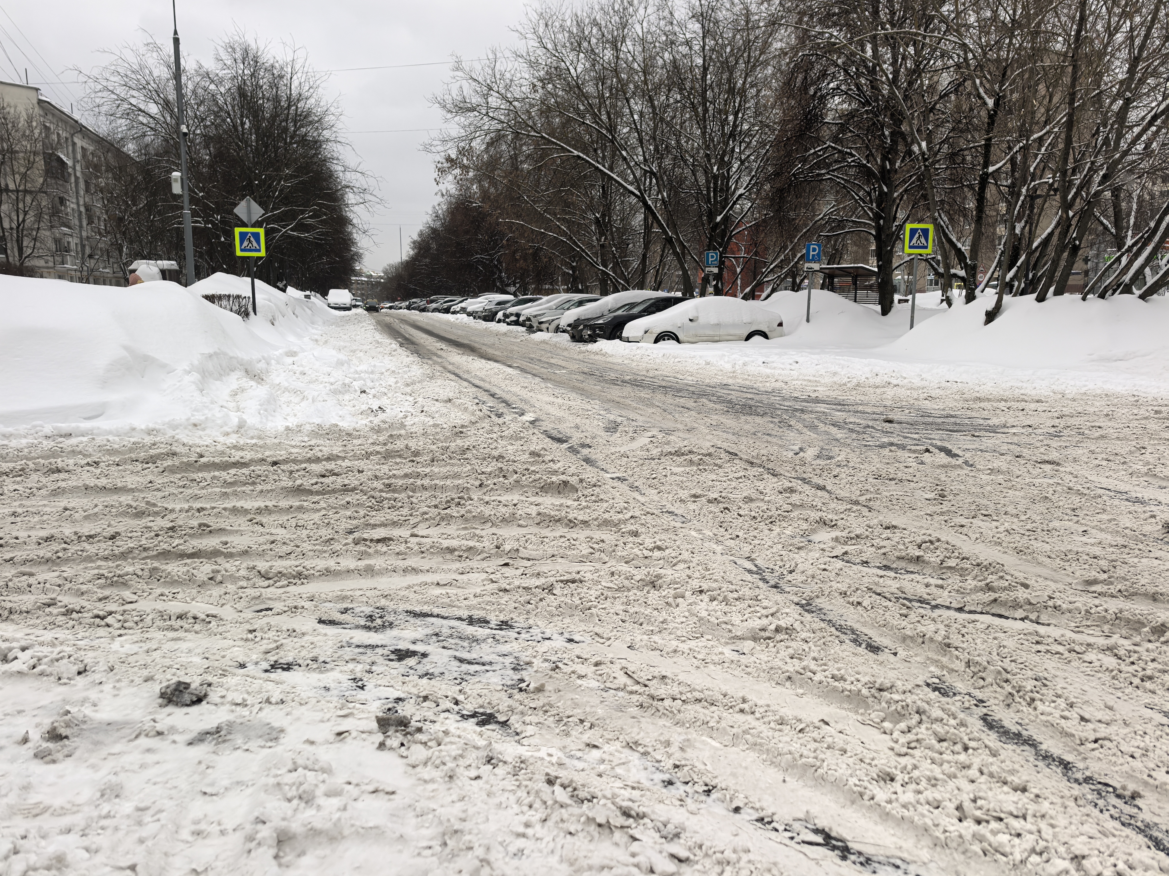 Участок городской дороги с пешеходным переходом, обозначенным знаками, в зимних условиях. Дорога покрыта снегом и наледью, припаркованные автомобили стоят вдоль обочины, частично перекрывая обзор.