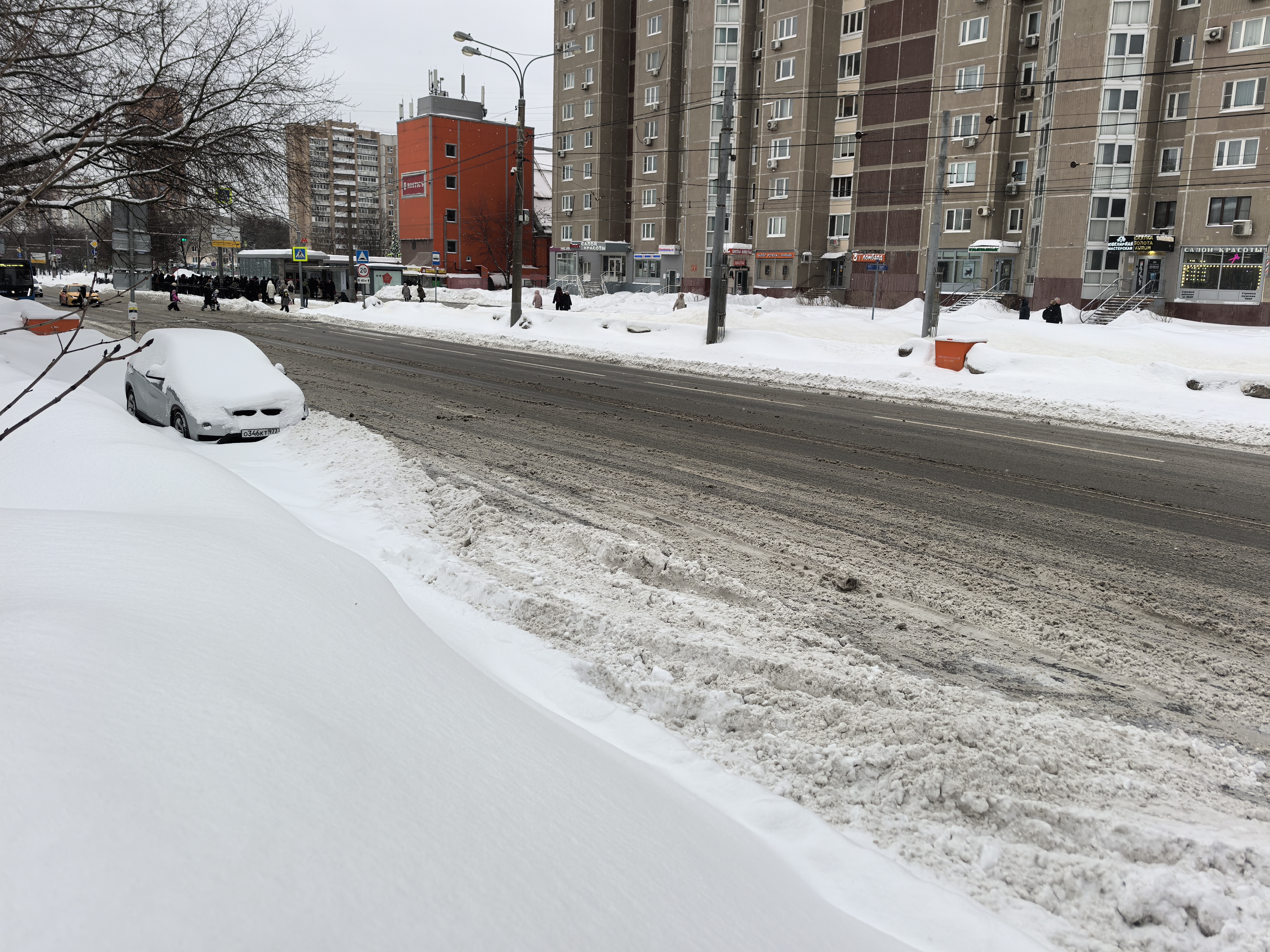 Участок городской дороги без пешеходного перехода в зимний период. Видны многоэтажные жилые дома, тротуары засыпаны снегом, проезжая часть очищена, но покрыта слякотью. На обочине припаркован автомобиль, частично скрытый снегом.