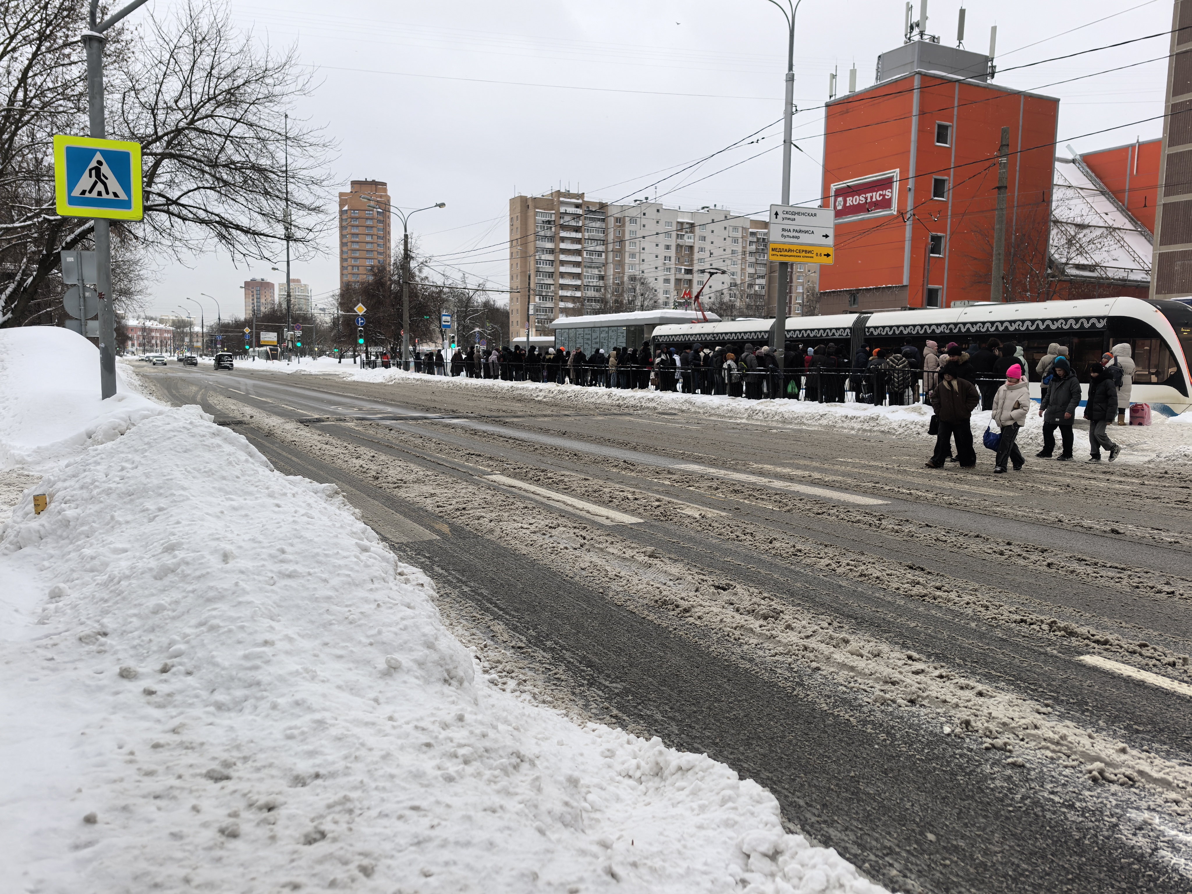Городская проезжая часть с пешеходным переходом, оборудованным знаками и разметкой. На обочине скопления людей, вероятно, у остановки общественного транспорта.