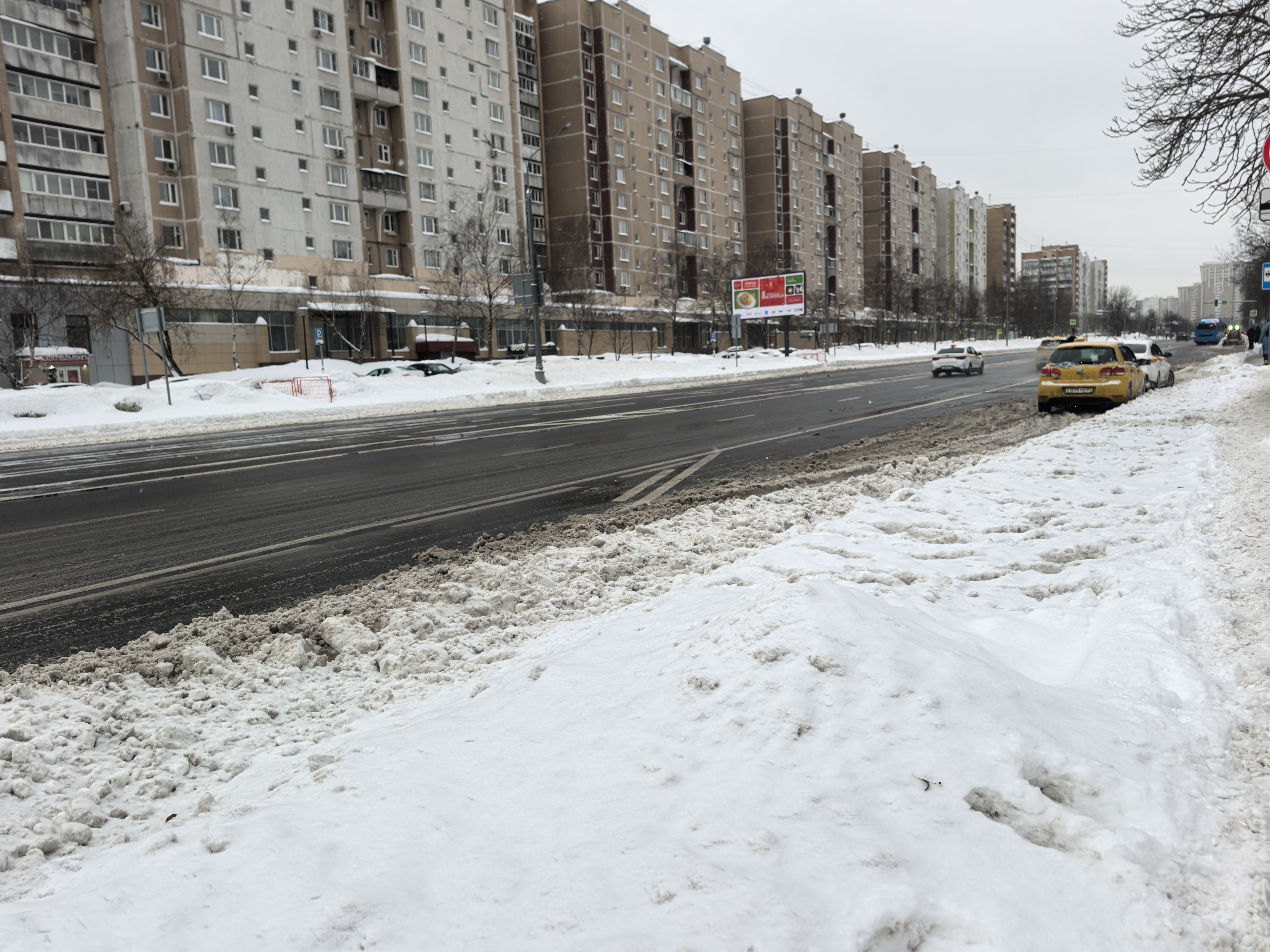 Широкая городская проезжая часть с многоэтажными домами, тротуары засыпаны снегом, пешеходный переход в кадре отсутствует.