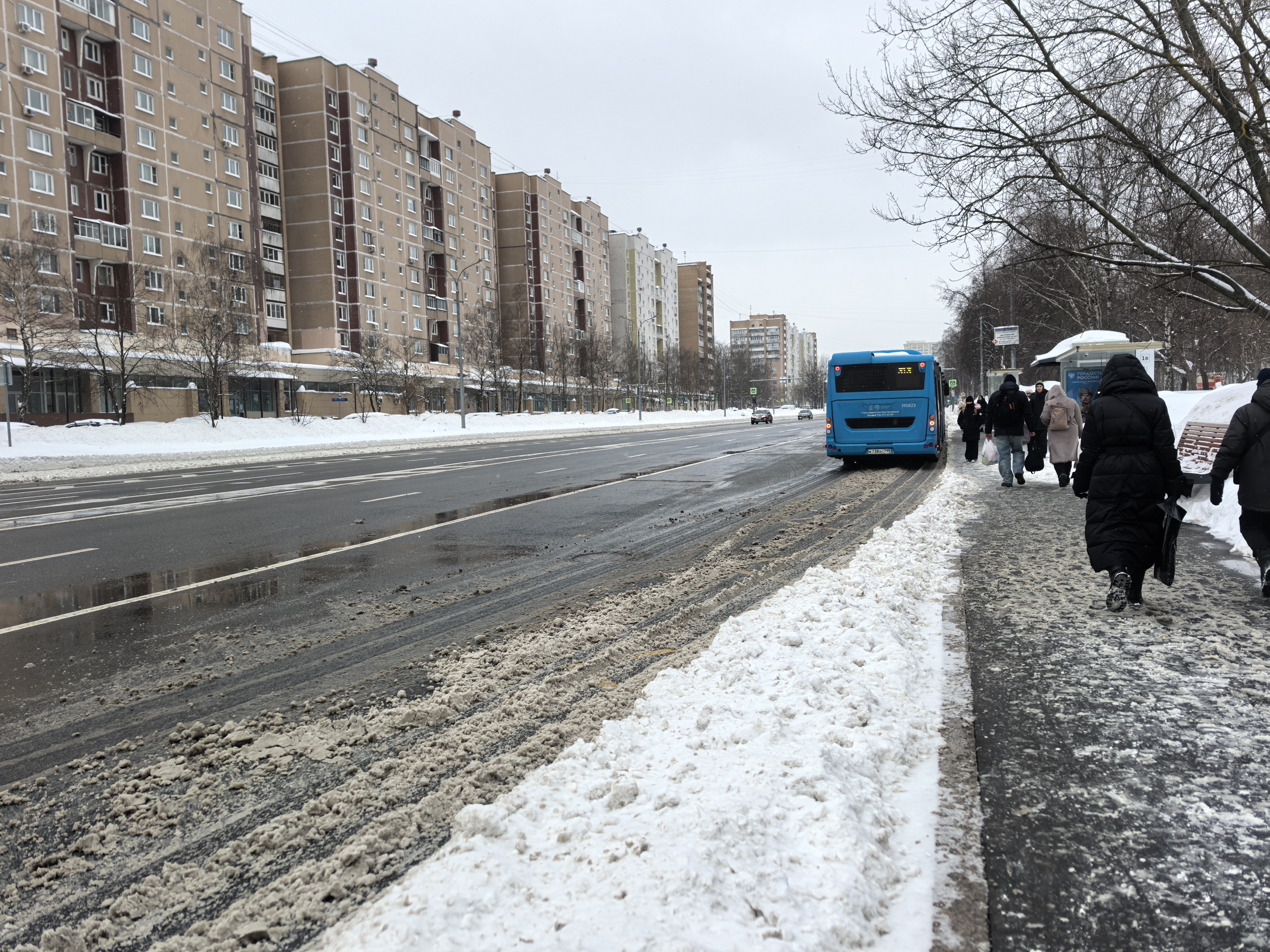 Участок городской улицы с широкой проезжей частью и тротуаром. Пешеходный переход в кадре отсутствует, виден только остановочный павильон. Дорога мокрая, с заметным слоем снежной шуги на обочине.