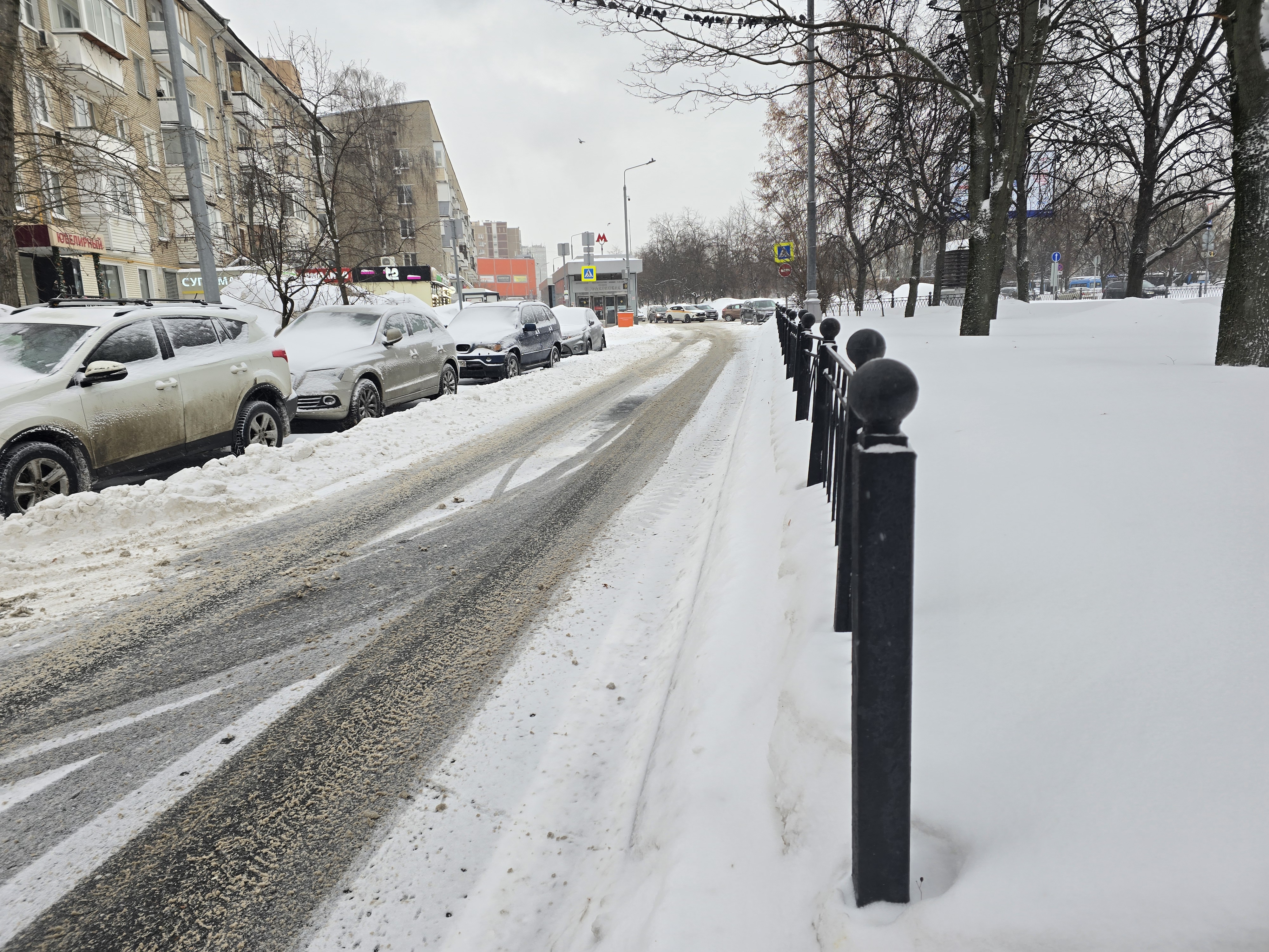 Участок городской улицы с припаркованными автомобилями и пешеходной зоной, отделенной забором. Пешеходный переход виден вдалеке (знак), но на данном участке его нет.