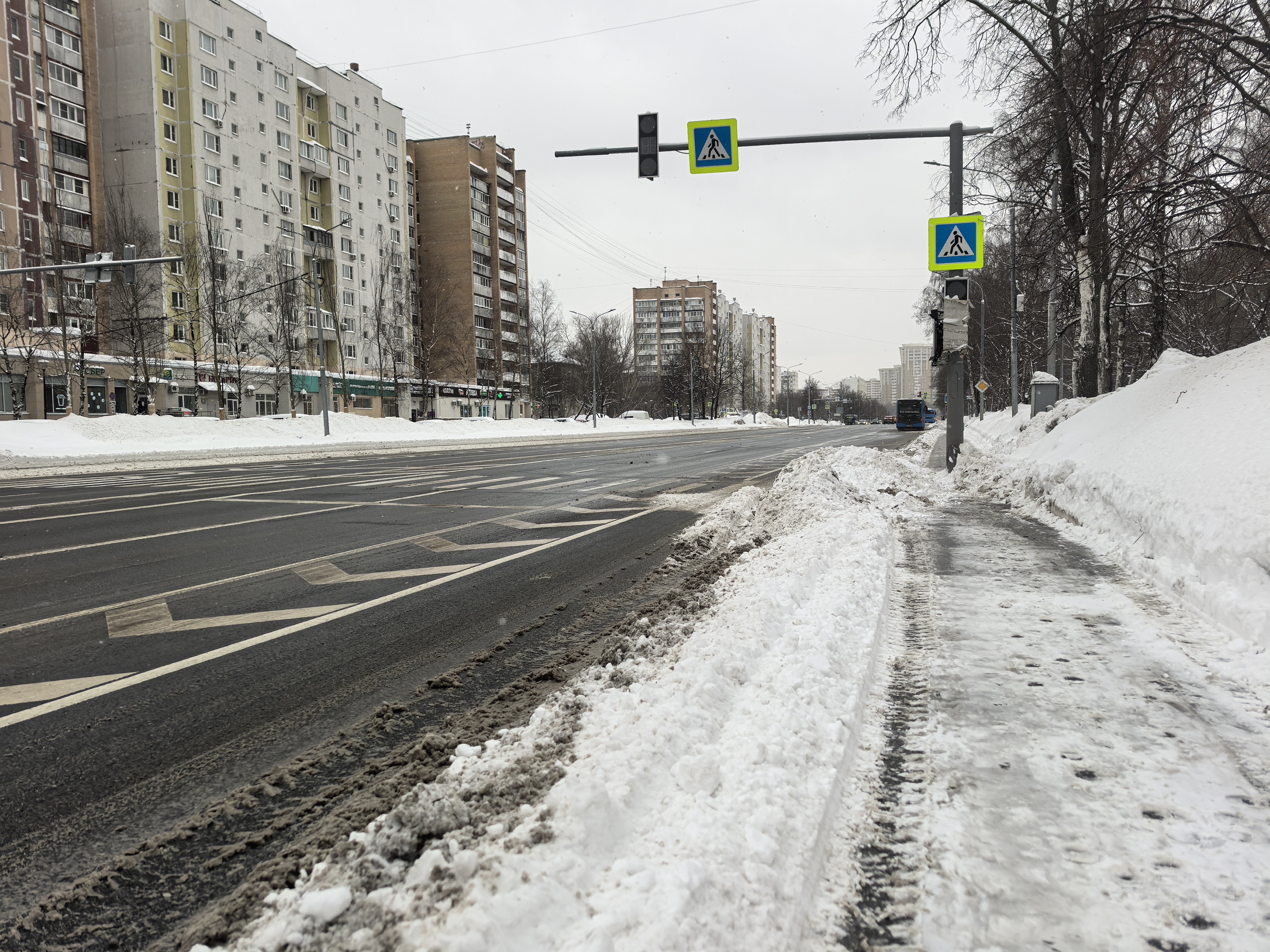 Широкая городская улица с пешеходным переходом, регулируемым светофором. Снежный покров на обочинах и тротуаре, проезжая часть очищена.