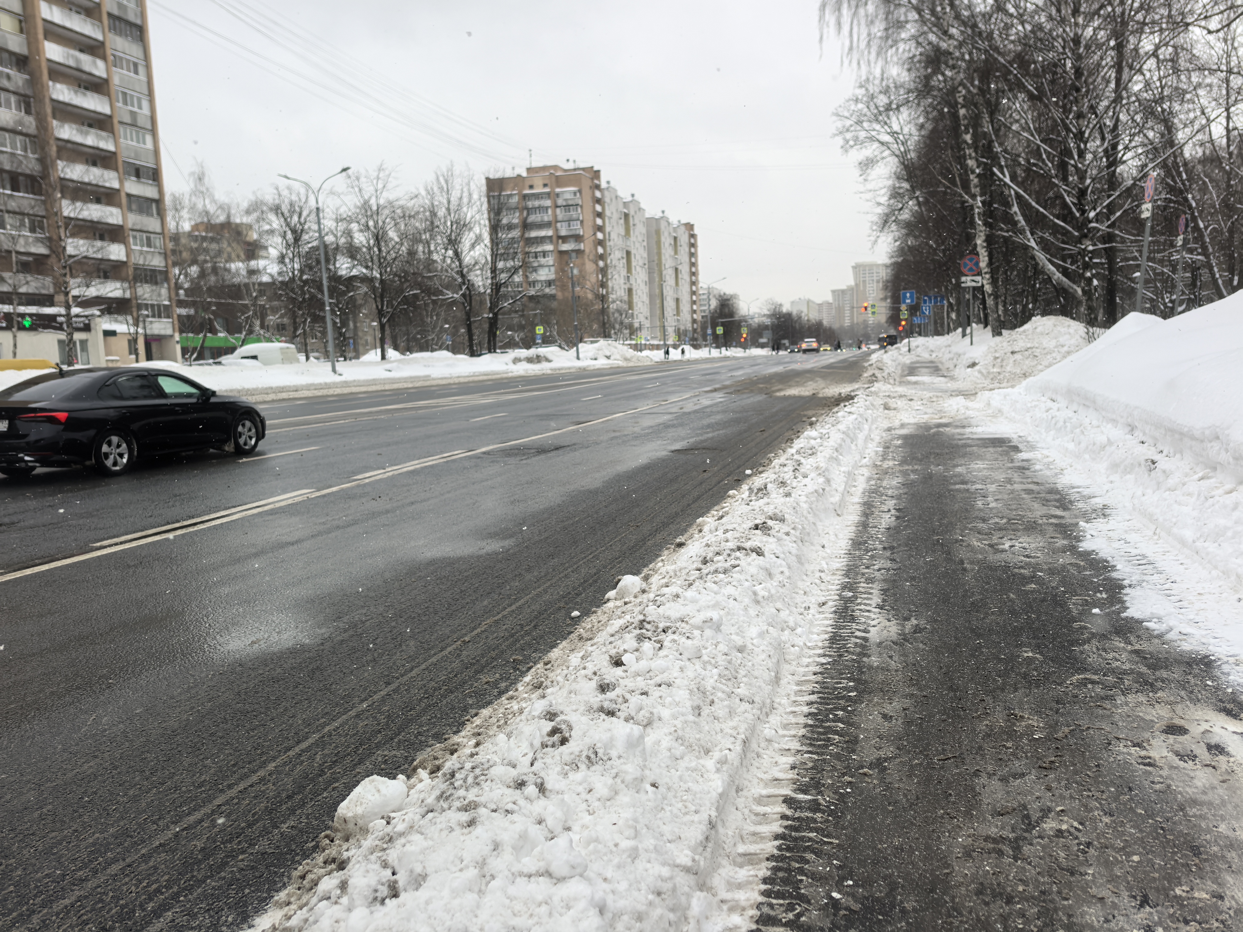 Широкая городская улица с многоэтажными домами, тротуаром, покрытым снегом и льдом, и проезжей частью с движущимся транспортом. Пешеходный переход в кадре отсутствует.