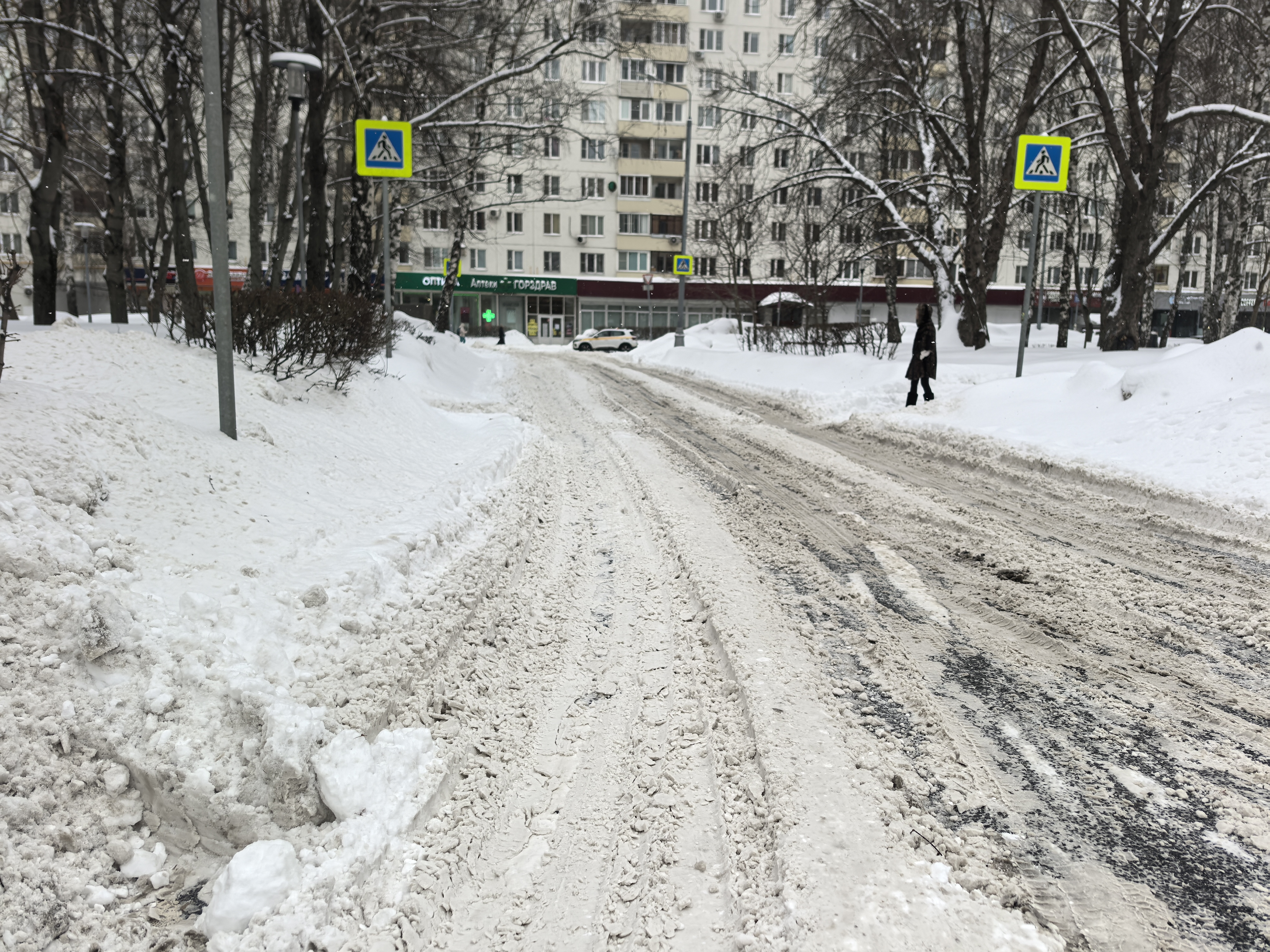 Участок городской дороги с пешеходным переходом в зимний период. На проезжей части видны следы от шин, дорога покрыта снегом и слякотью. Пешеходный переход обозначен знаками, но сама зебра полностью скрыта под снегом.
