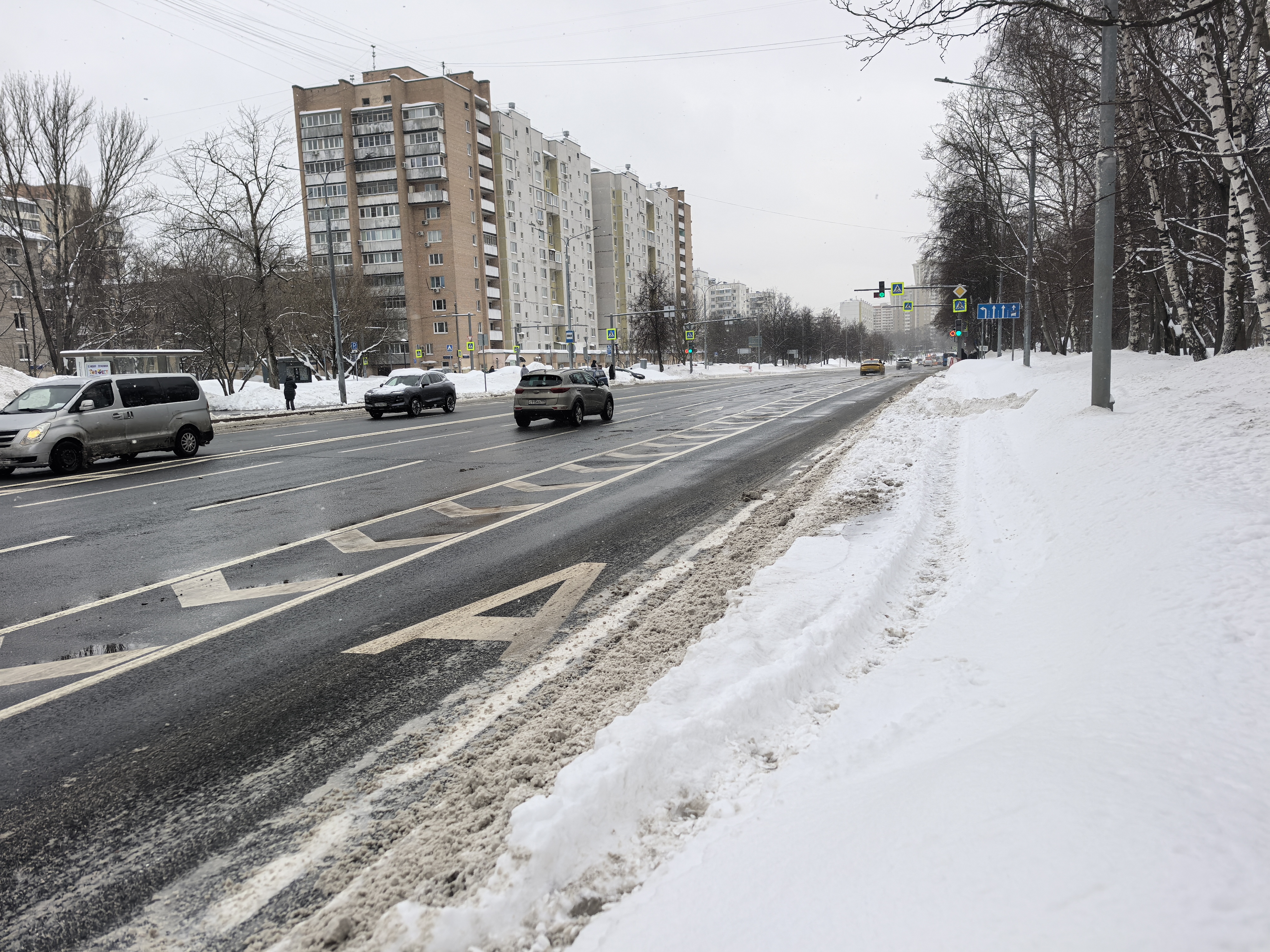 Широкая городская улица с пешеходным переходом, регулируемым светофорами. Видны проезжающие автомобили и припаркованный фургон на обочине.