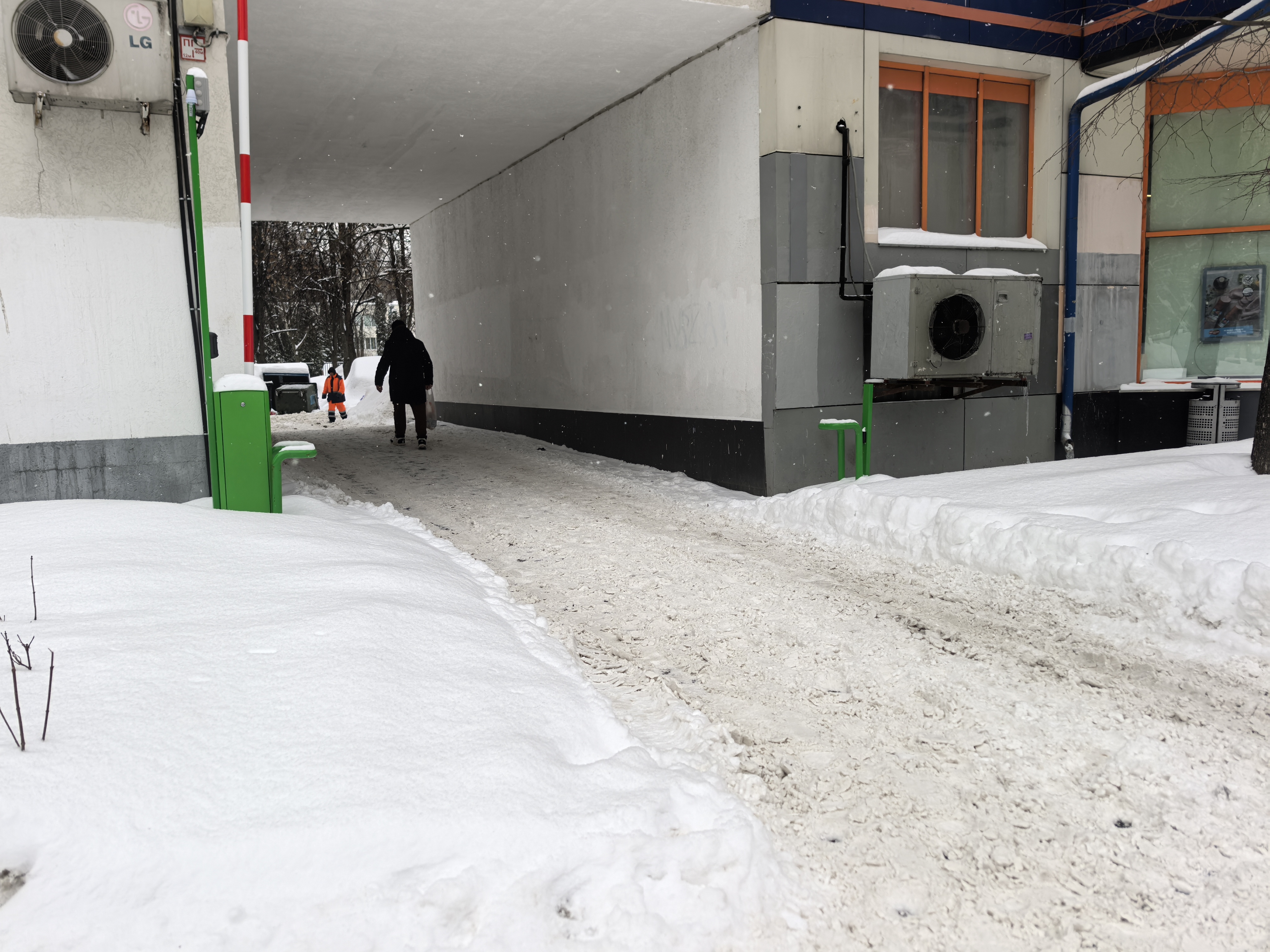 Дворовая территория, проход под навесом здания. Видны следы от шин на снегу, что указывает на возможность проезда транспорта, но пешеходы передвигаются по очищенной от снега дорожке.