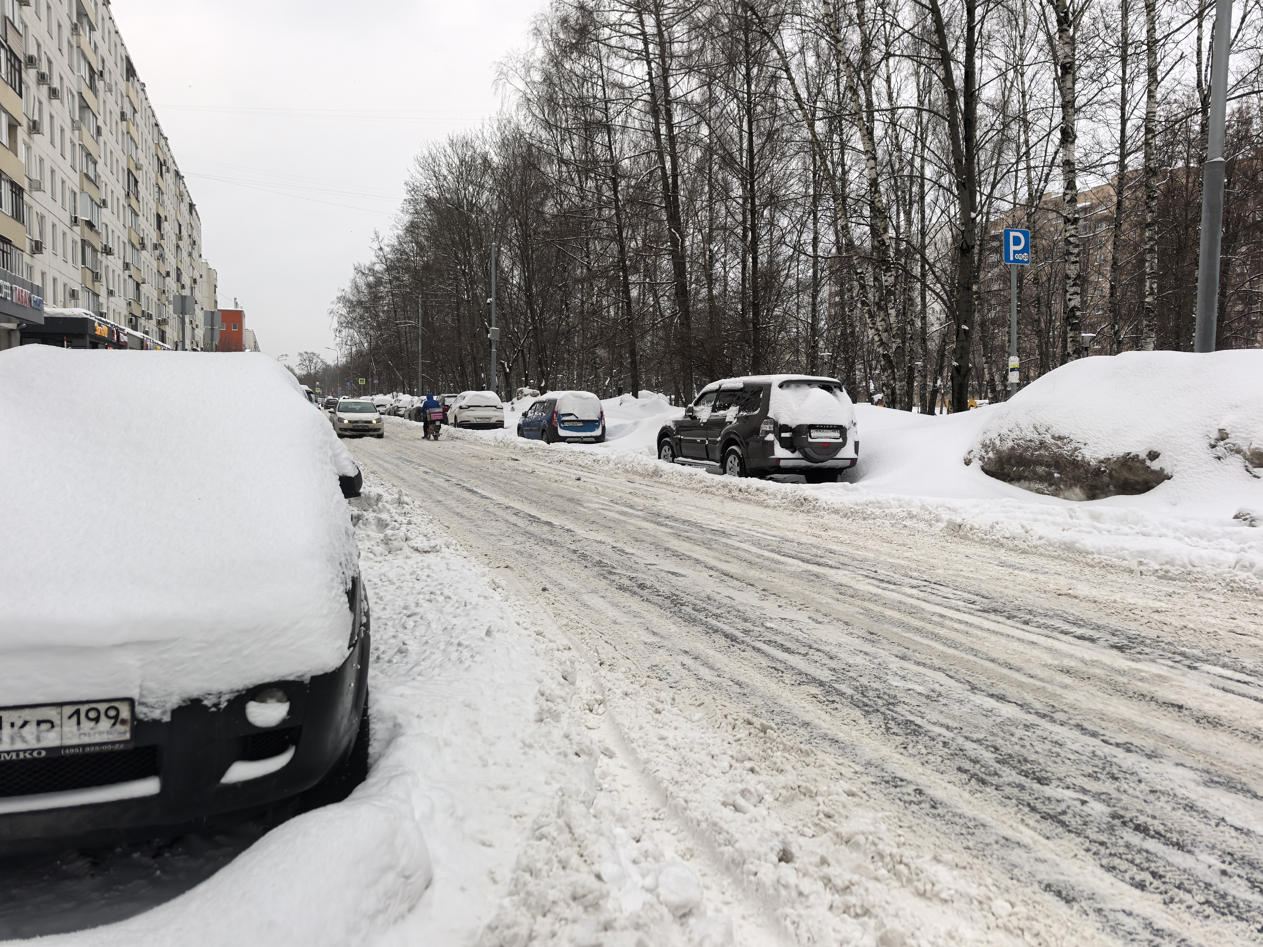 Городская улица в зимний период, покрытая снегом и ледяной коркой. Тротуары полностью засыпаны снегом, проезжая часть узкая из-за припаркованных автомобилей.