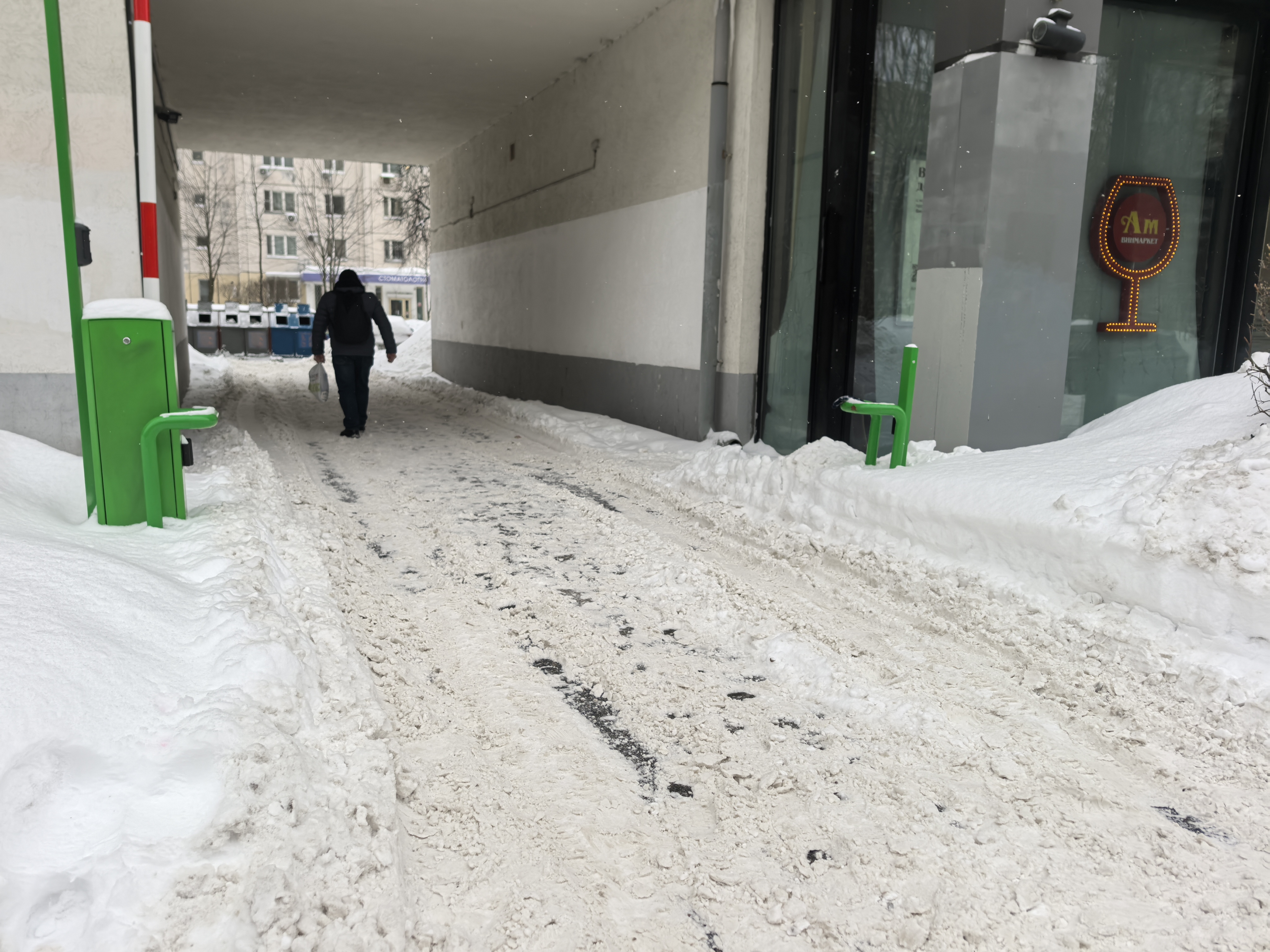 Дворовая территория, проход под зданием (проход к мусорным бакам). Дорога покрыта снегом и ледяной кашей, видны следы от колес. Присутствуют зеленые столбики-ограничители.