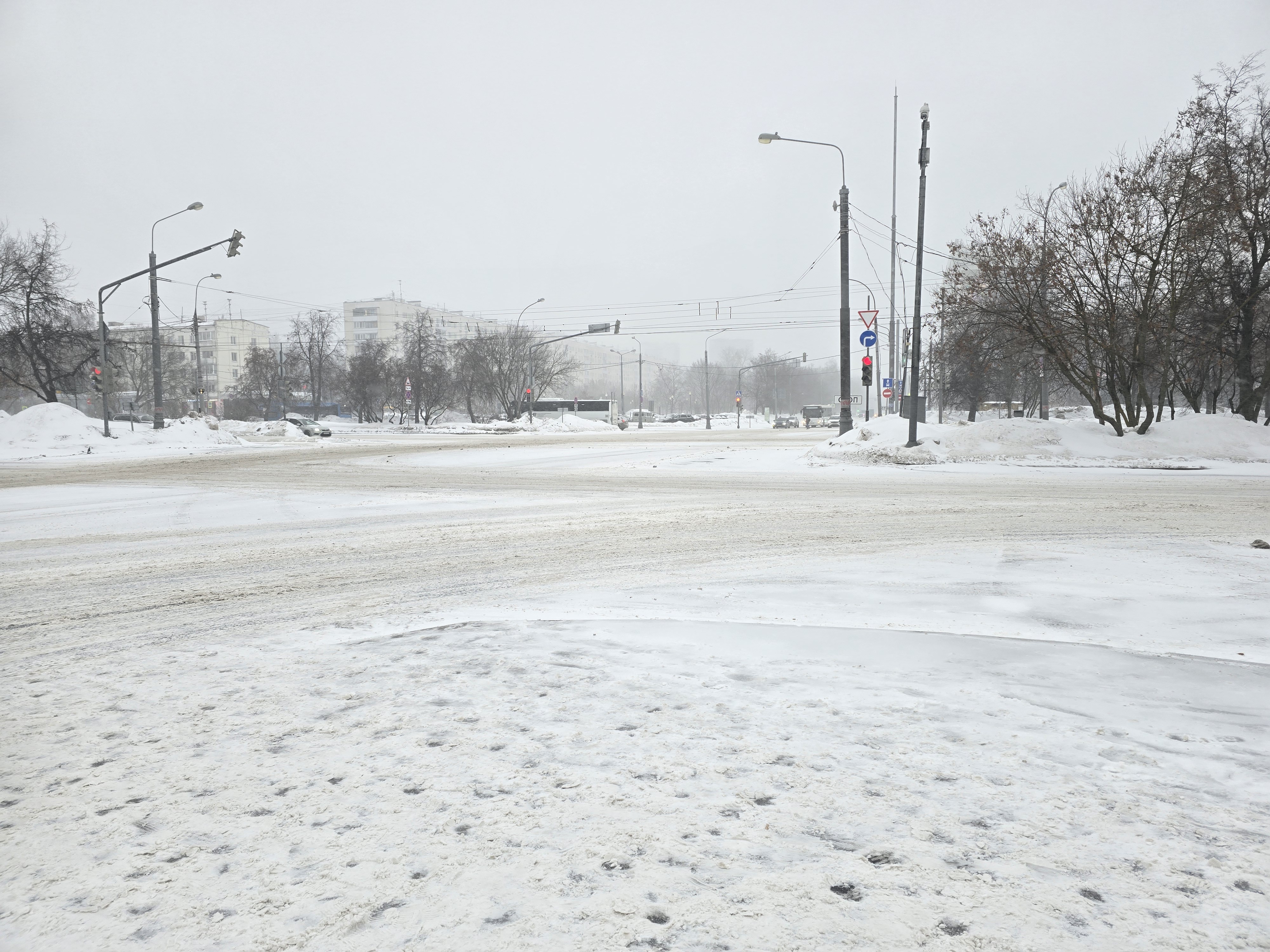 Широкая проезжая часть с широким тротуаром, полностью покрытым снегом. Пешеходный переход не виден в кадре, видны только дорожные знаки и светофоры на перекрестке.