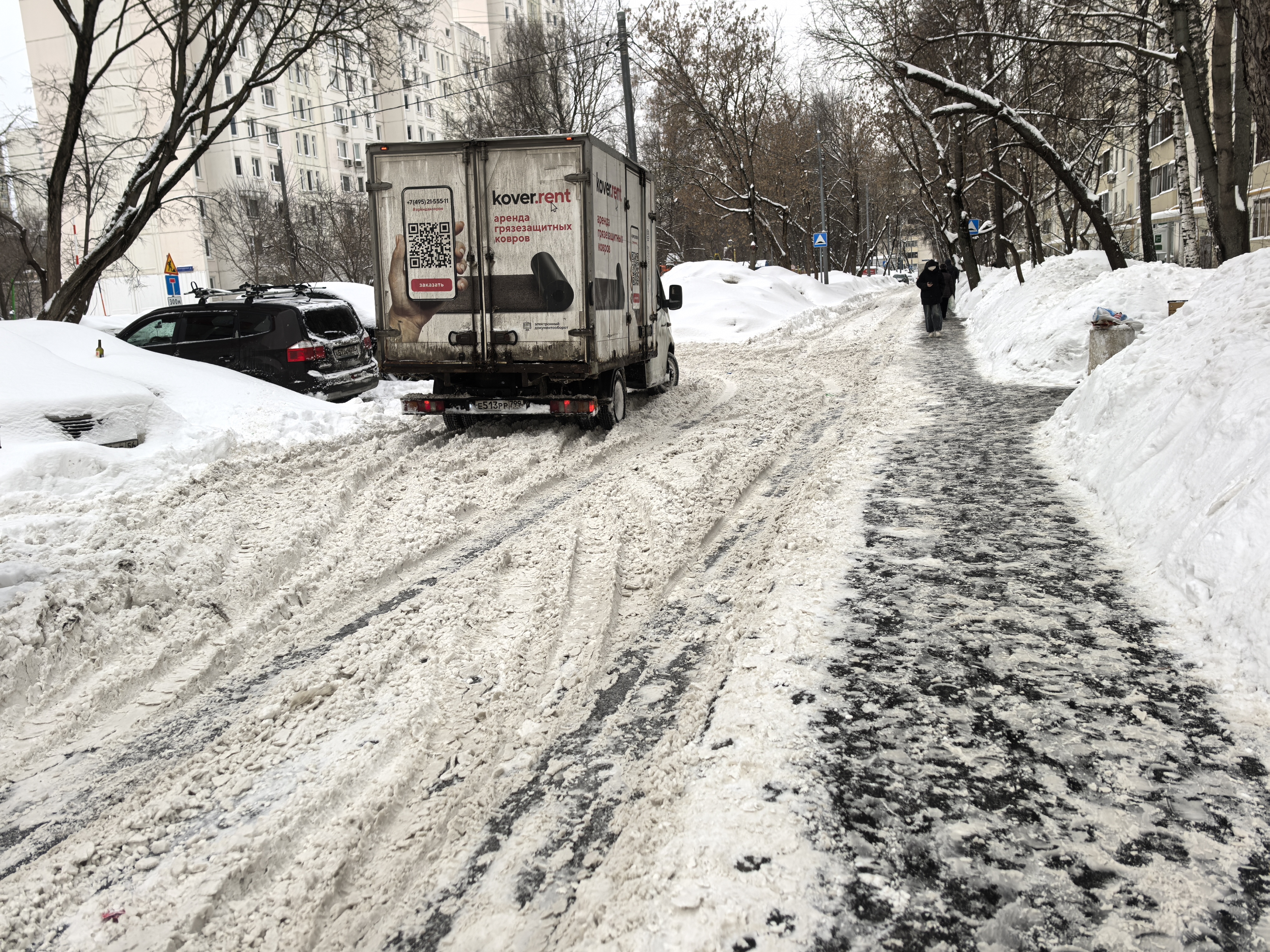 Участок городской улицы зимой без пешеходного перехода. Тротуар очищен, но проезжая часть покрыта снегом и льдом. Видны припаркованные автомобили и движущийся грузовик.