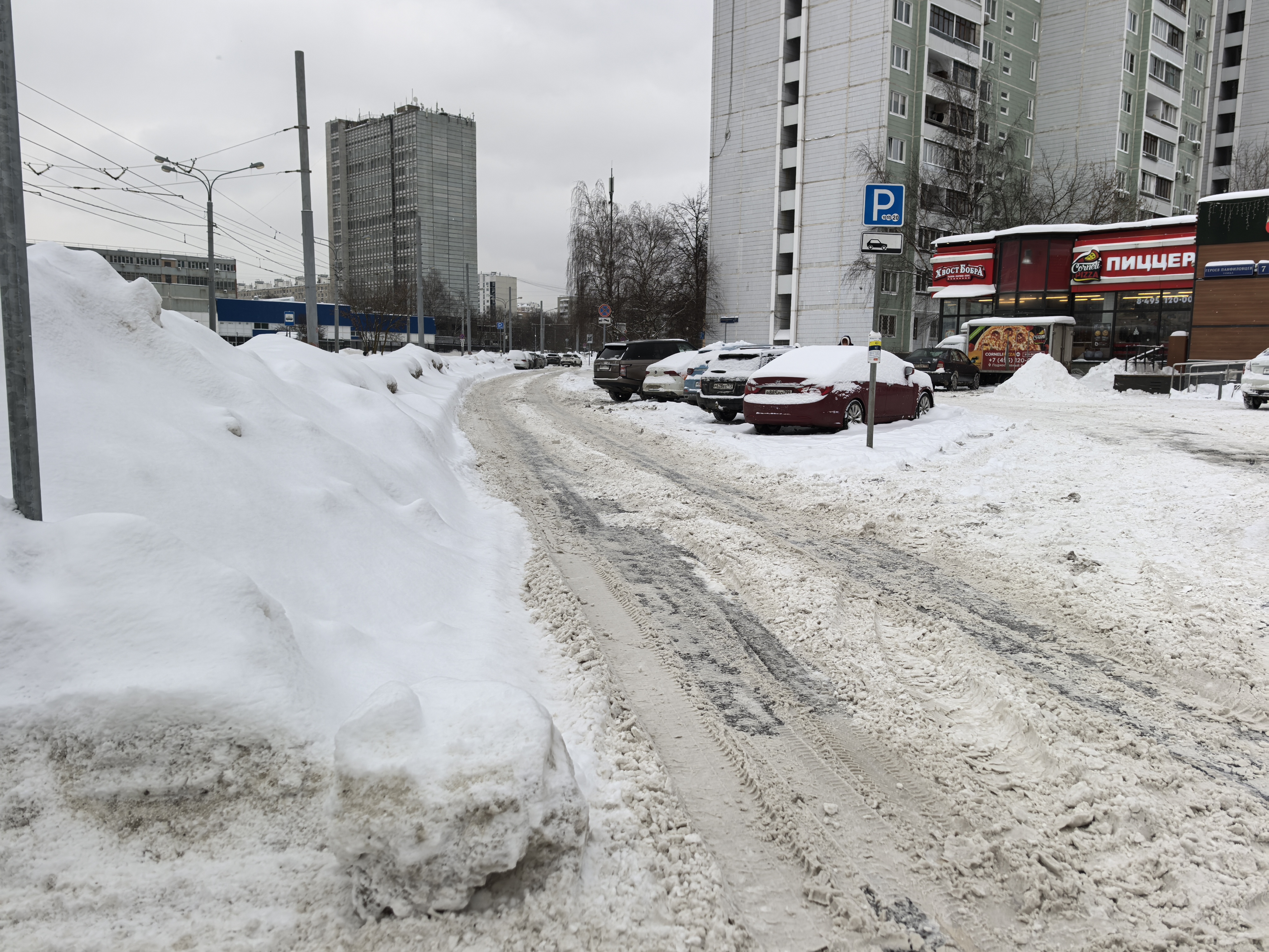 Участок проезжей части без обустроенного пешеходного перехода, покрытый снегом и ледяной коркой. Справа припаркованы автомобили, слева — высокие сугробы.