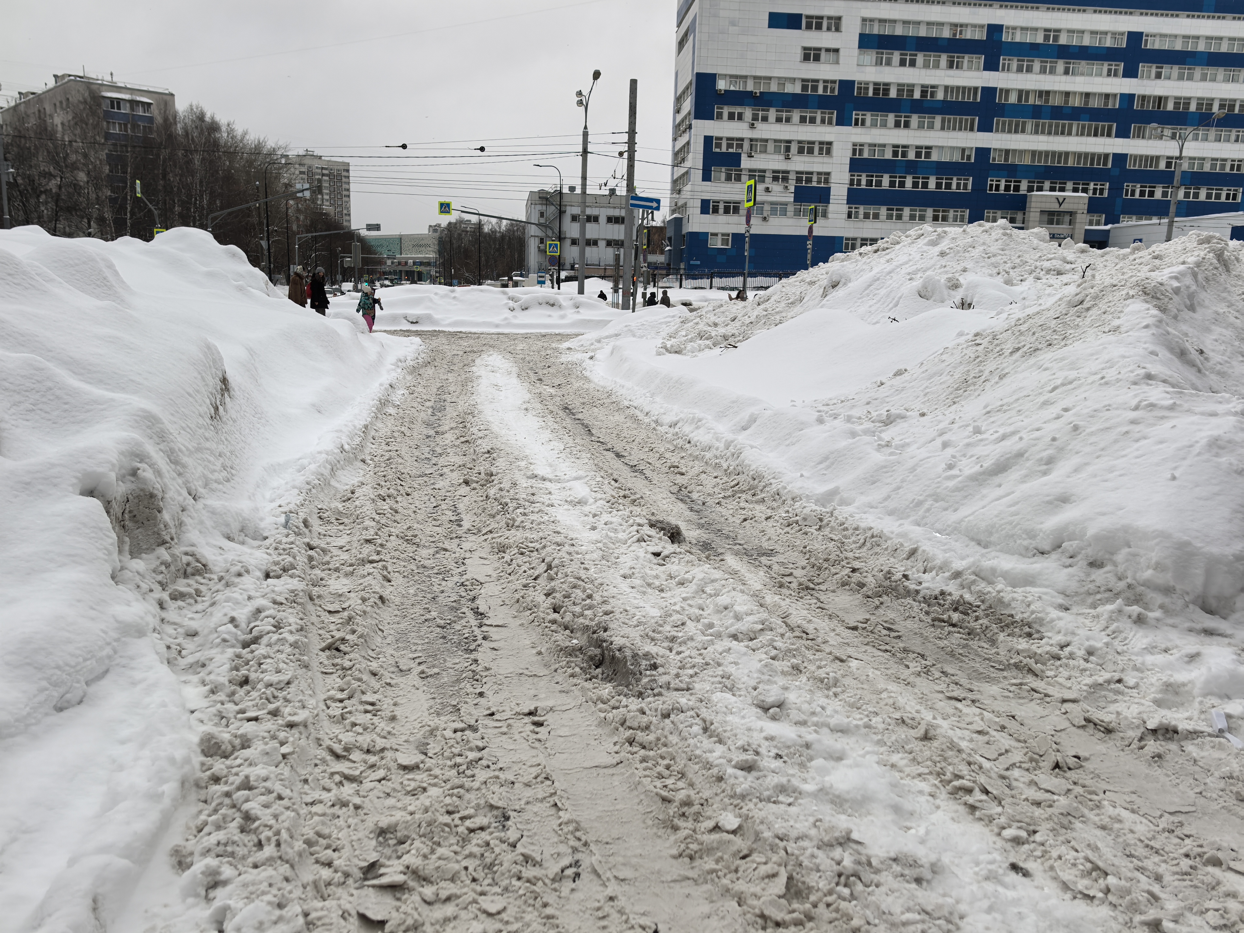 Участок дороги без пешеходного перехода, с высокой насыпью снега по бокам, на проезжей части видны следы от шин и снежно-грязевая каша. На заднем плане виден знак пешеходного перехода, но сам переход не в зоне кадра.