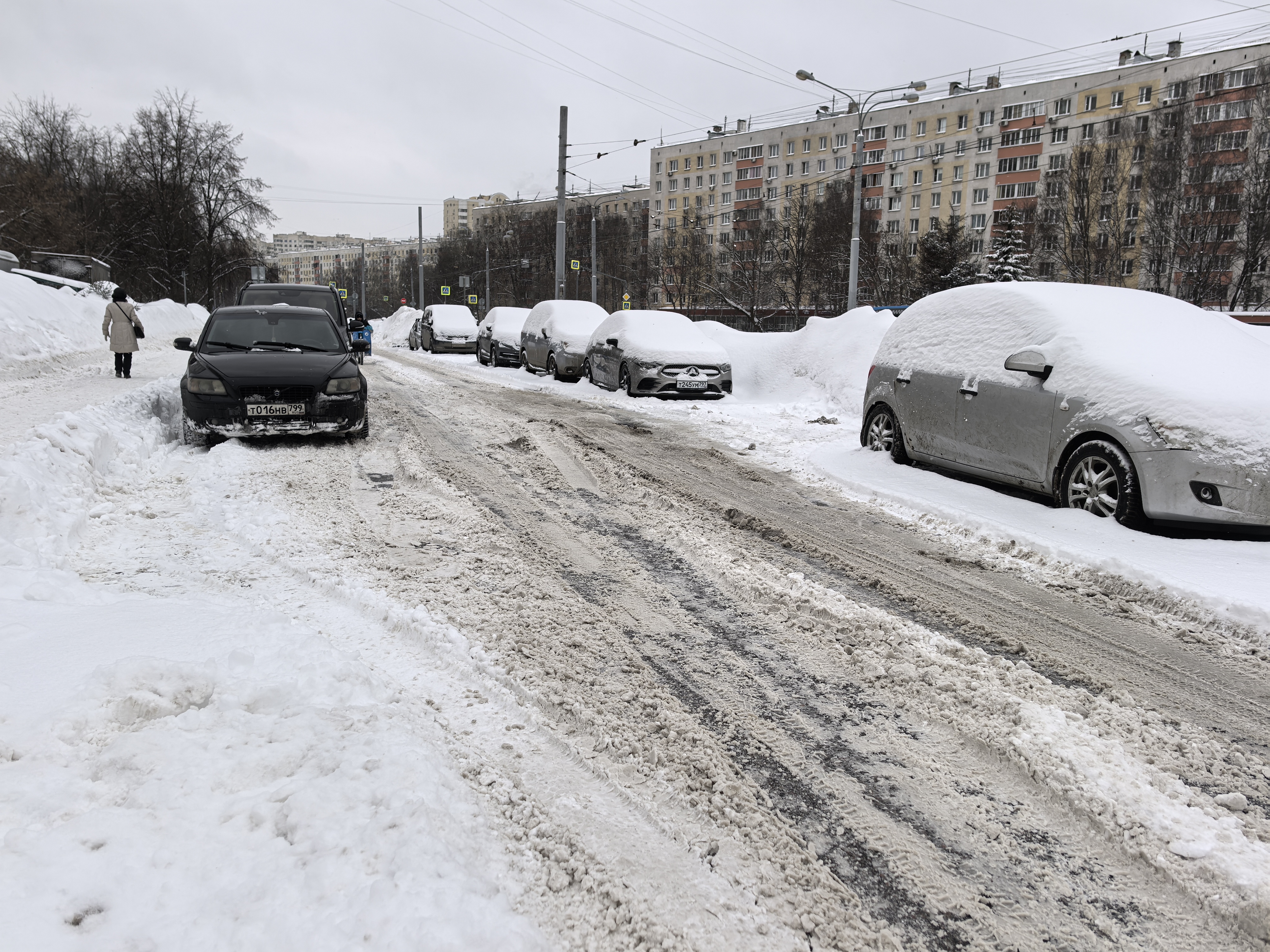 Участок городской дороги без обустроенного пешеходного перехода. Дорожное покрытие полностью покрыто снегом и слякотью, видны колеи. С обеих сторон дороги припаркованы автомобили, частично занесённые снегом. На заднем плане видны жилые дома и знаки пешехо