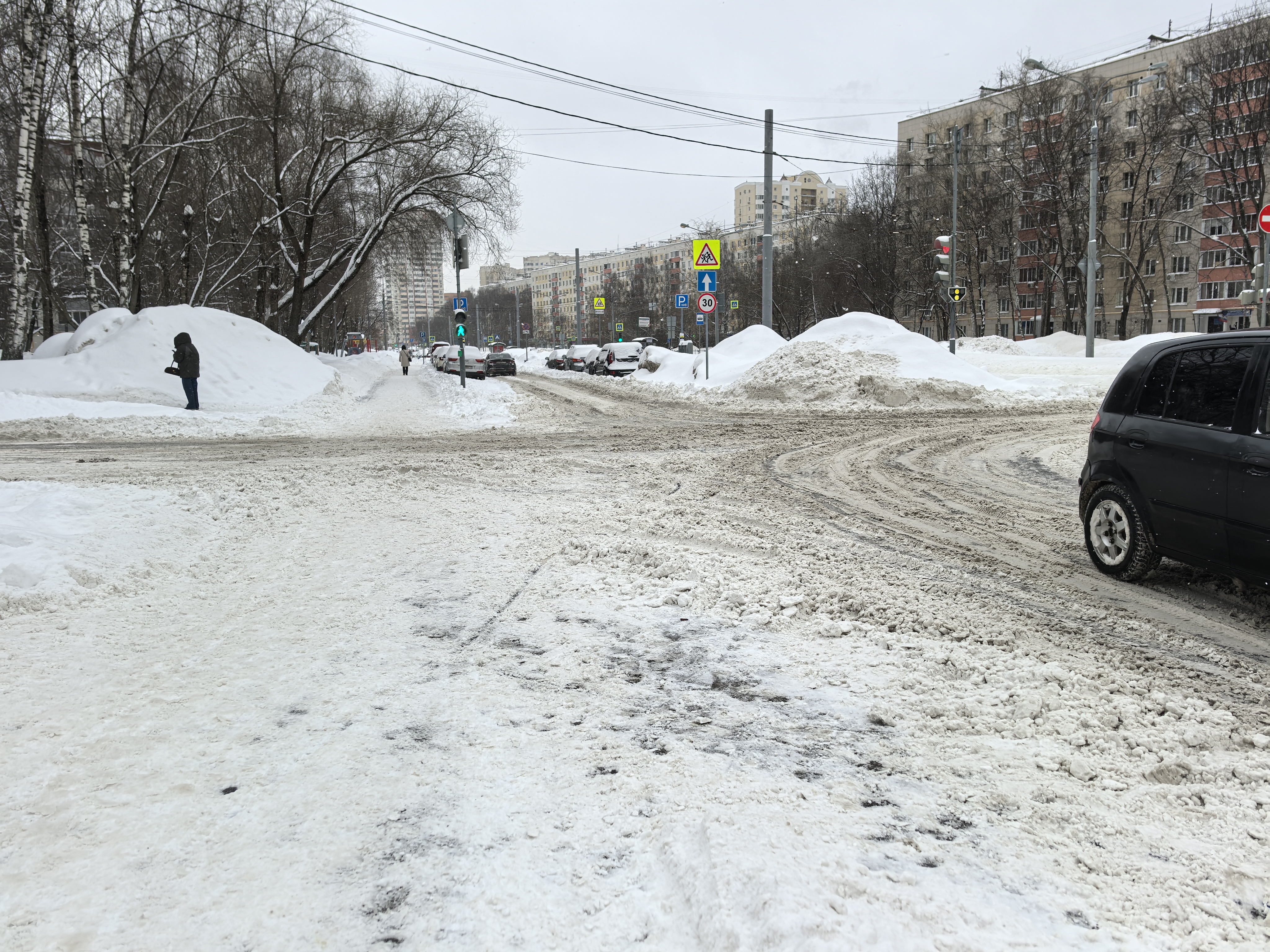 Городская улица с пешеходным переходом (зебра), регулируемым светофором, в зимних условиях. Видны припаркованные автомобили вдоль дороги и большие сугробы по бокам.