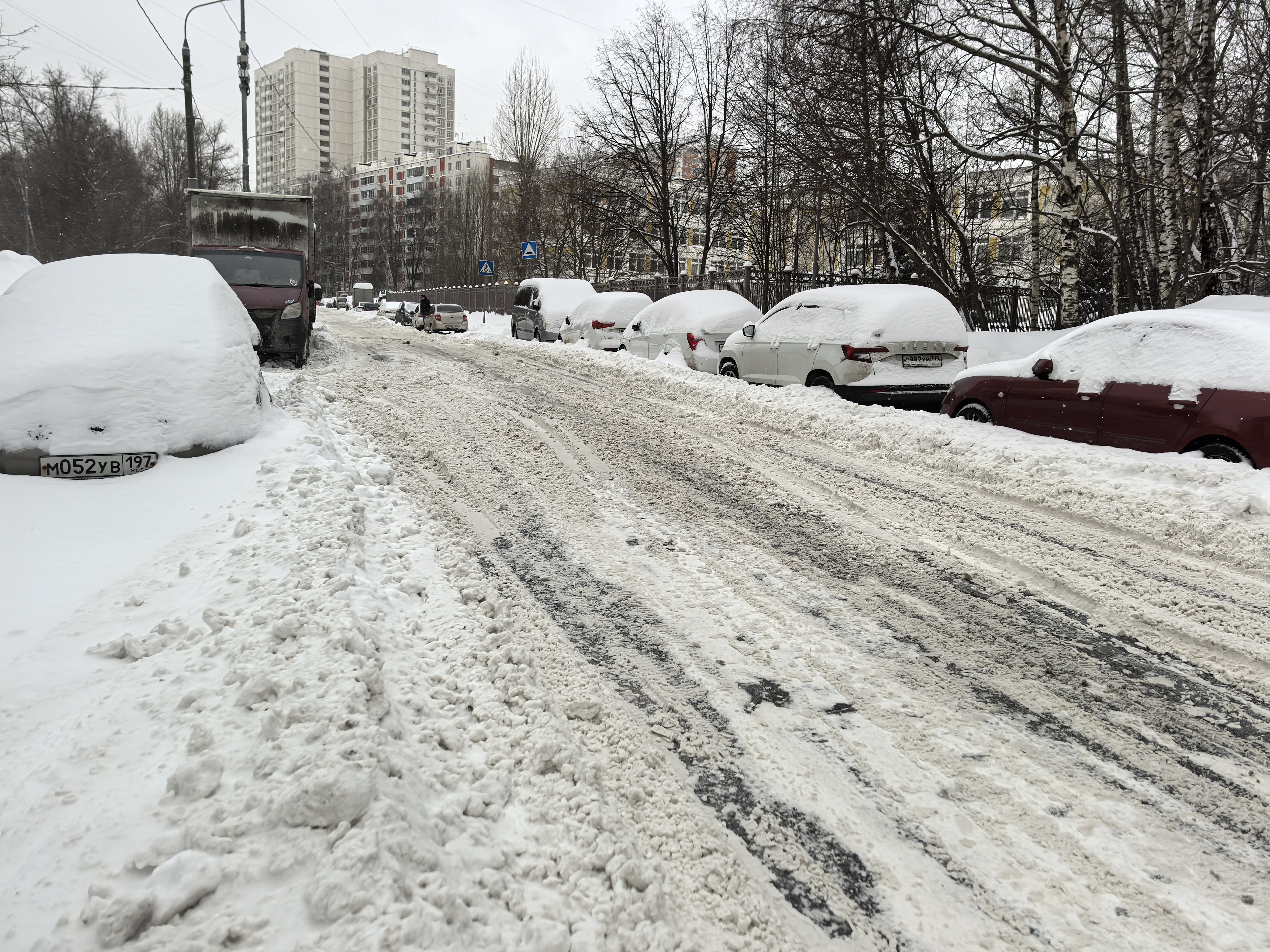 Проезжая часть в жилом районе, покрытая снегом и наледью. Пешеходного перехода (зебры) в видимой части нет, но есть знак «Пешеходный переход» впереди. Тротуары полностью заблокированы снегом.