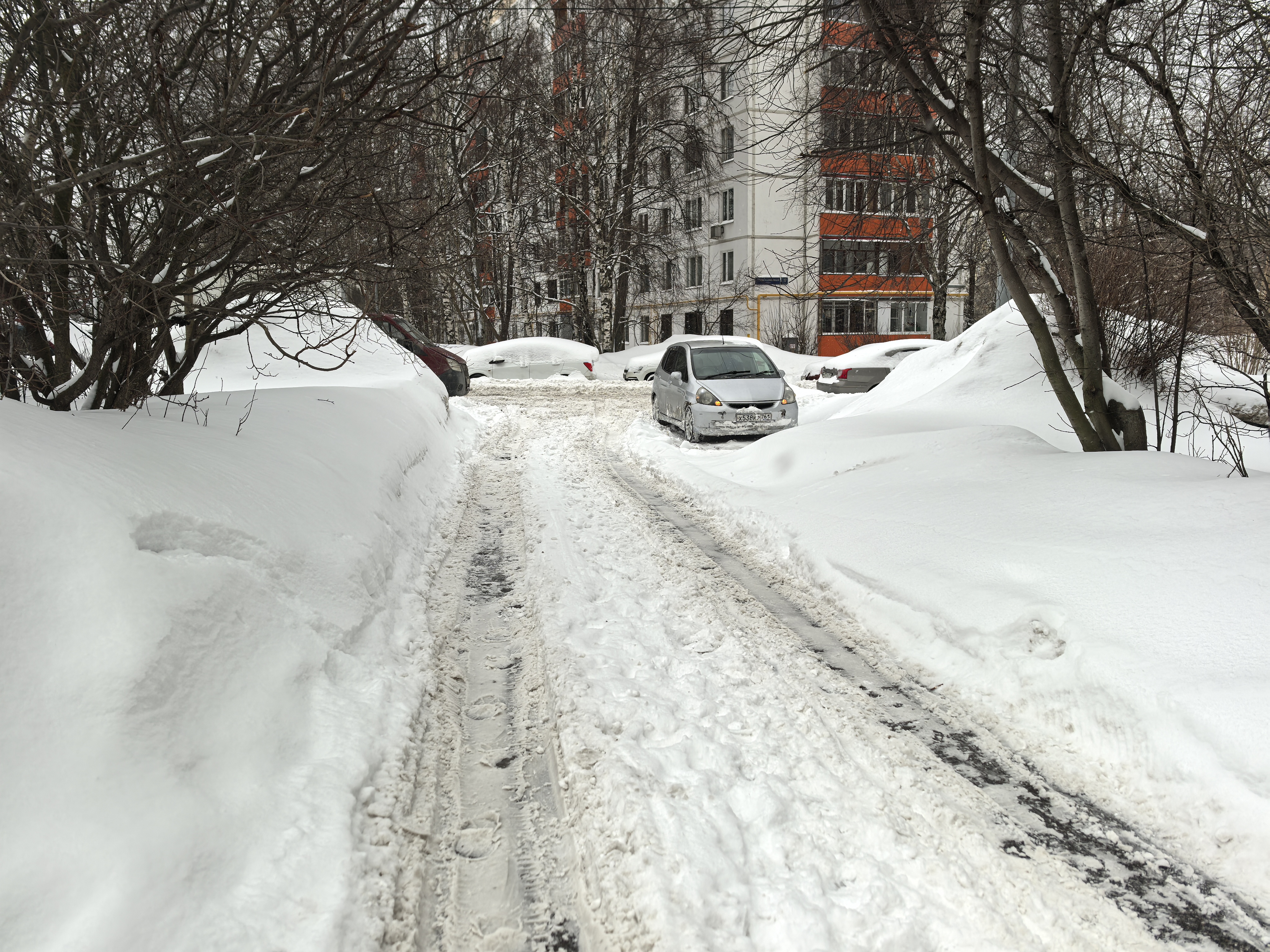 Дворовая территория, заснеженная проезжая часть между жилыми домами, припаркованные автомобили, отсутствуют тротуары и пешеходные переходы.