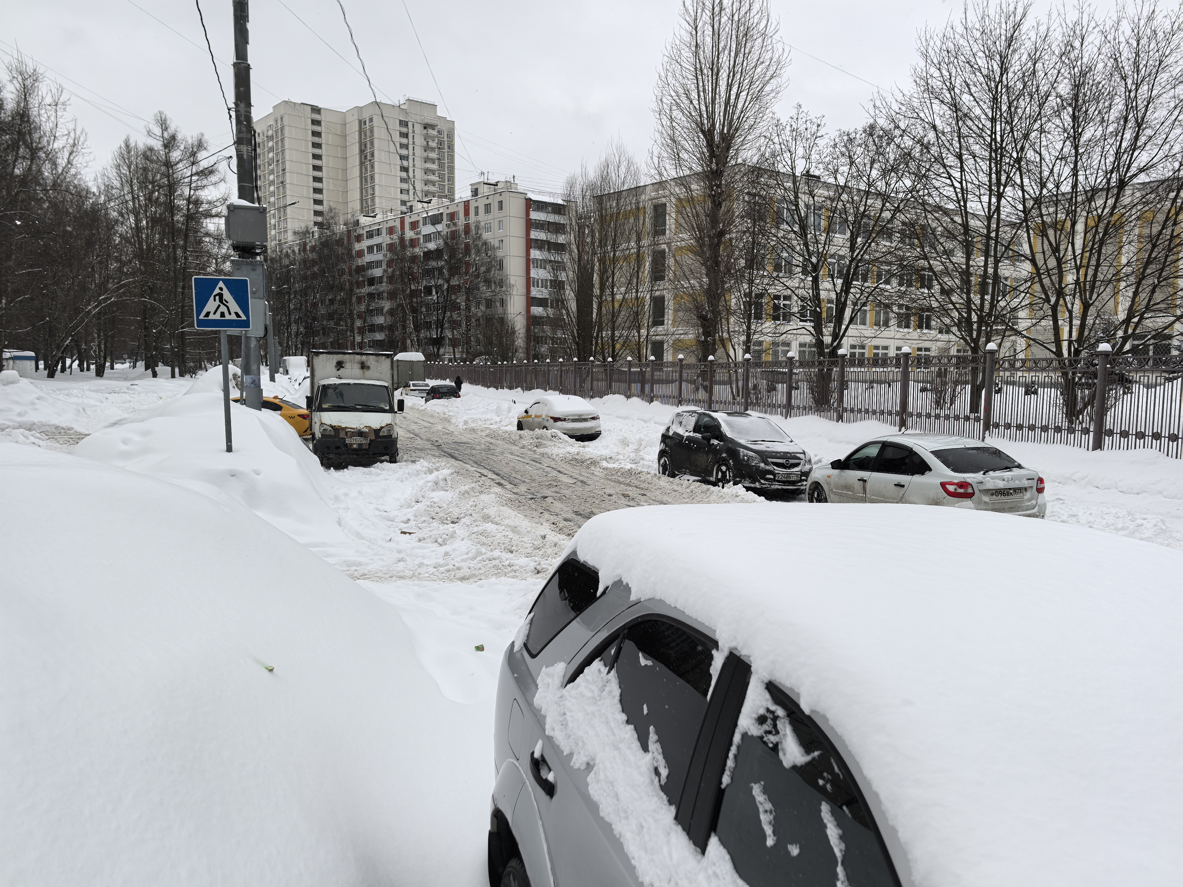 Участок проезжей части без видимого пешеходного перехода, с припаркованными автомобилями и сильным снежным покровом. На заднем плане видны жилые дома и ограждение.