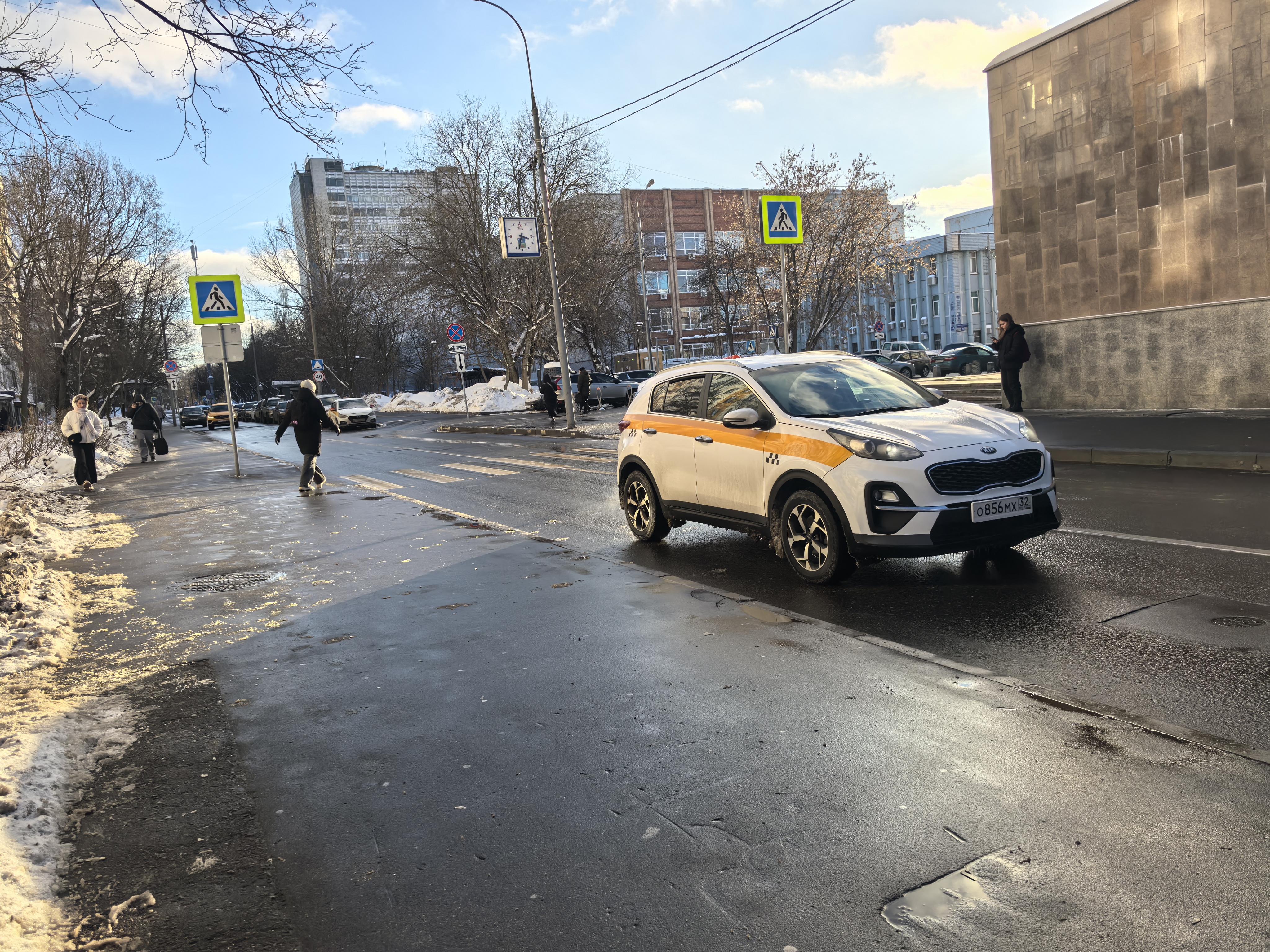 Городская улица с пешеходным переходом, обозначенным знаками и разметкой. На проезжей части движется автомобиль, тротуар частично покрыт снегом и грязью.