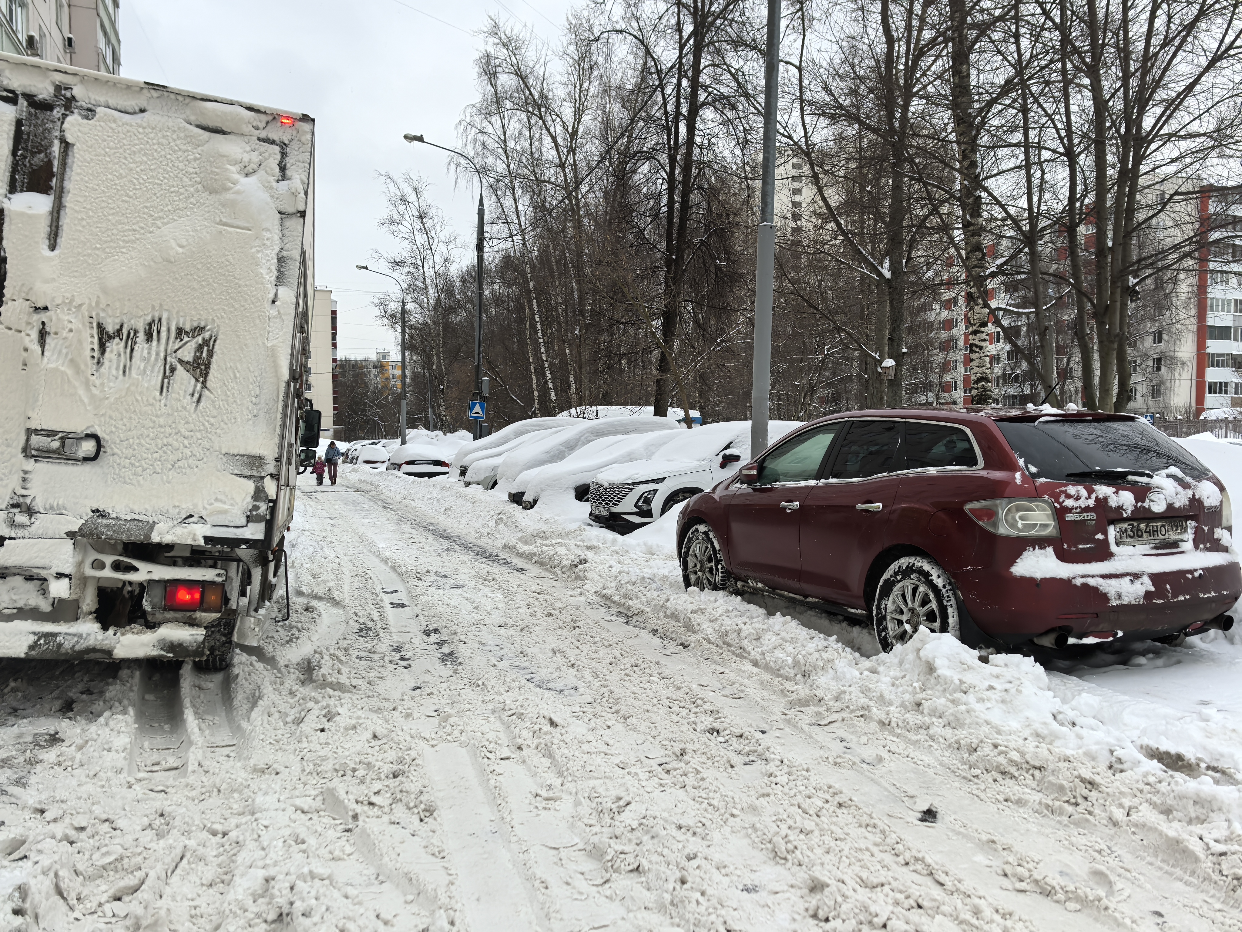 Проезжая часть в жилом районе, покрытая снегом и наледью. Тротуары не видны или заблокированы, пешеходы вынуждены идти по проезжей части. На обочине припаркованы автомобили, закрывающие обзор.