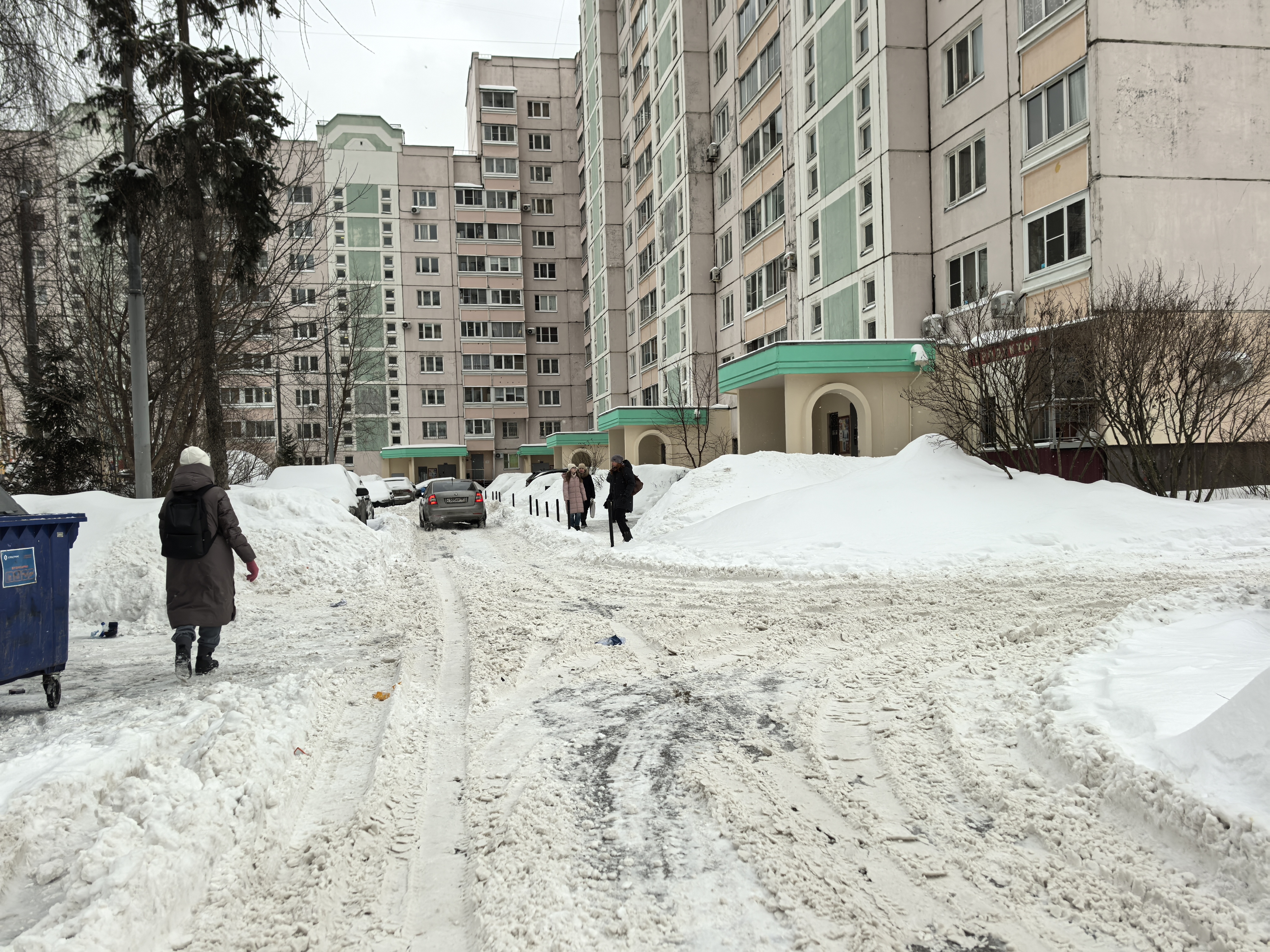 Дворовая территория между жилыми домами, покрытая снегом и колеей. Пешеходы идут по проезжей части, так как тротуары полностью засыпаны снегом.