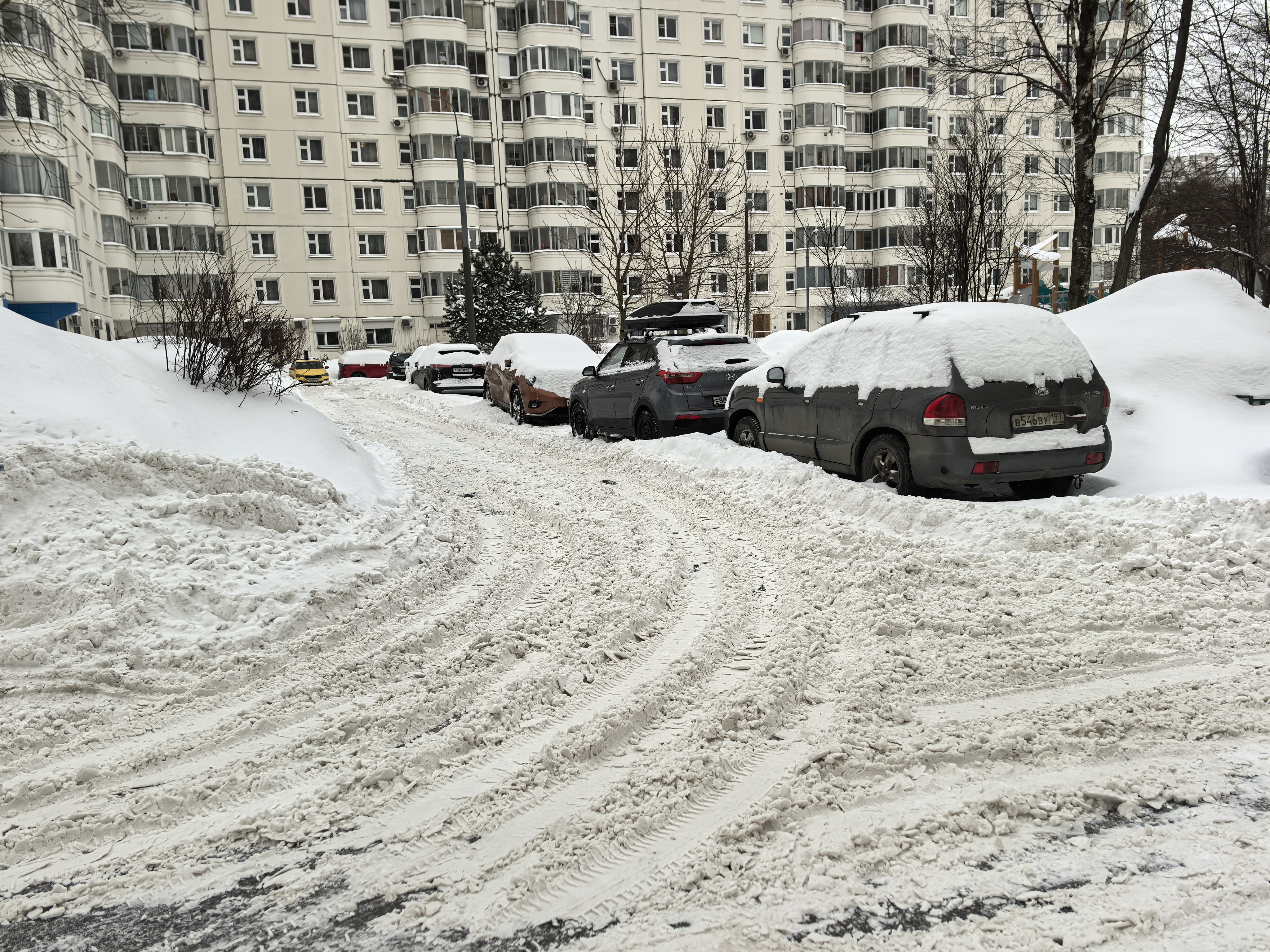 Дворовая территория с припаркованными автомобилями, покрытая снегом. Видны жилые дома и деревья. Дорога узкая, проход затруднен из-за припаркованных машин и снежных валов.