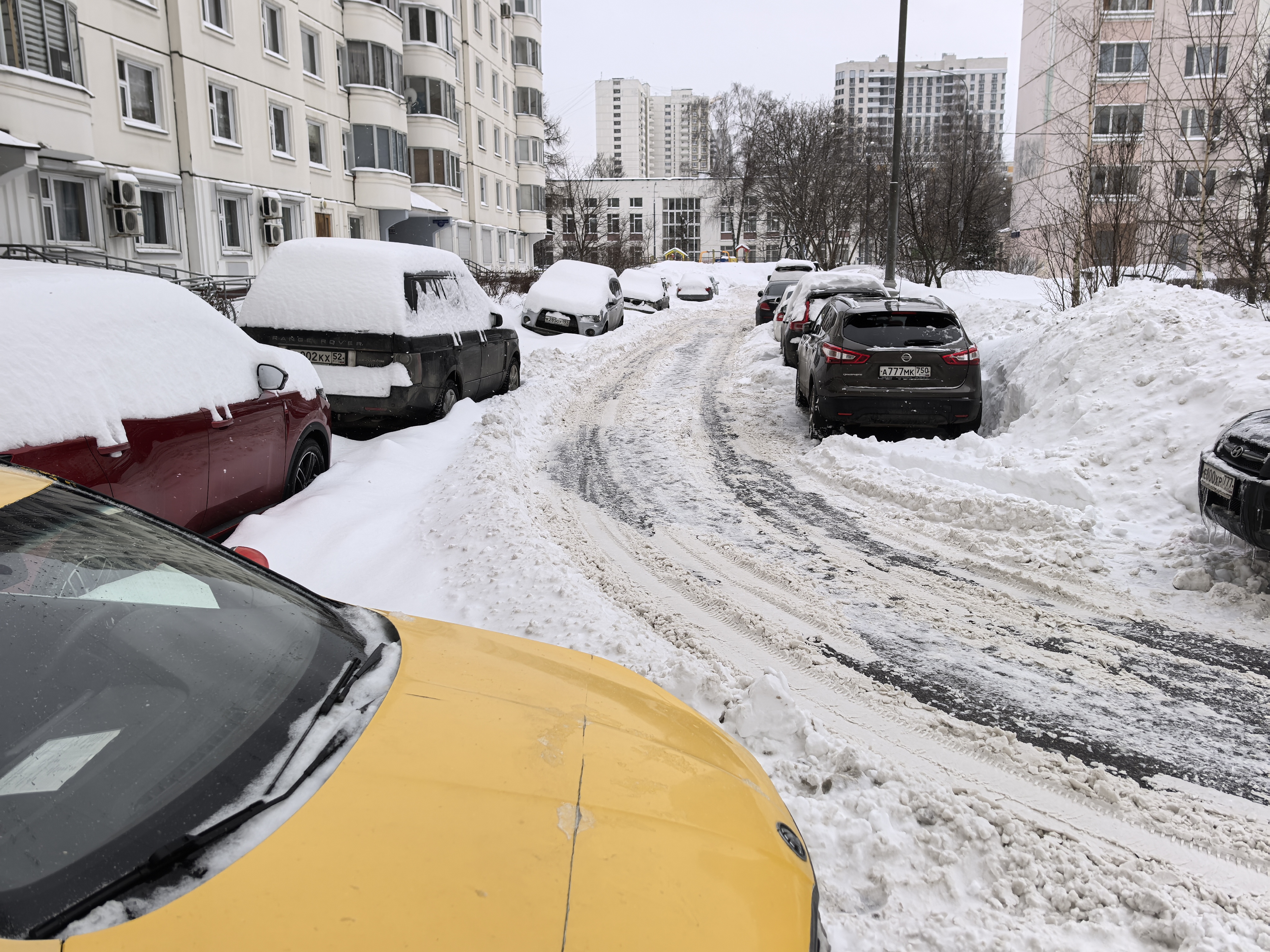 Дворовая территория с парковкой вдоль проезжей части, покрытой снегом. Проезжая часть узкая, заснеженная, с видимыми следами от шин. Припаркованные автомобили с обеих сторон сужают проезд.