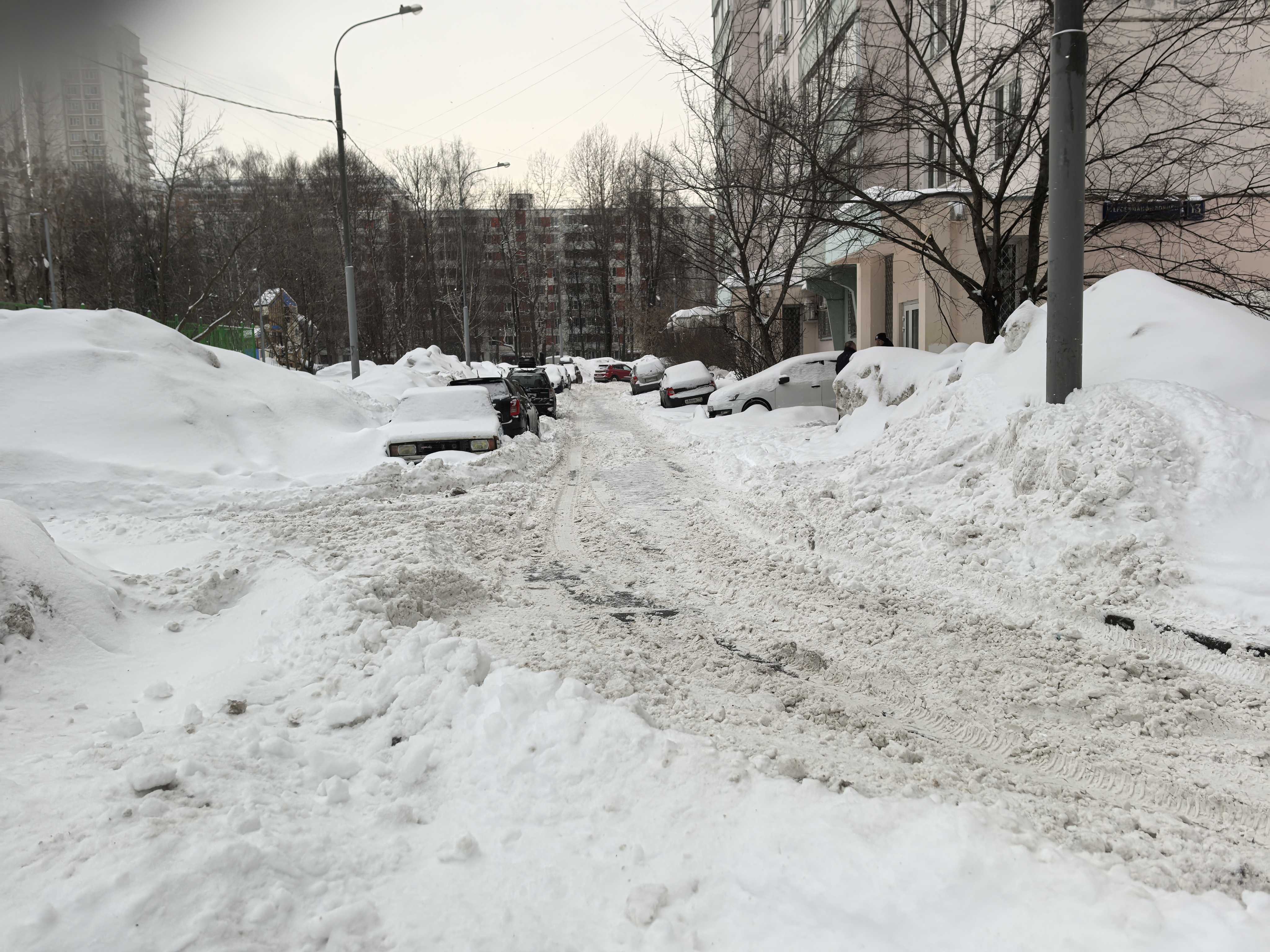 Дворовая территория с припаркованными автомобилями, покрытая снегом. Видны жилые дома и детская площадка на заднем плане.