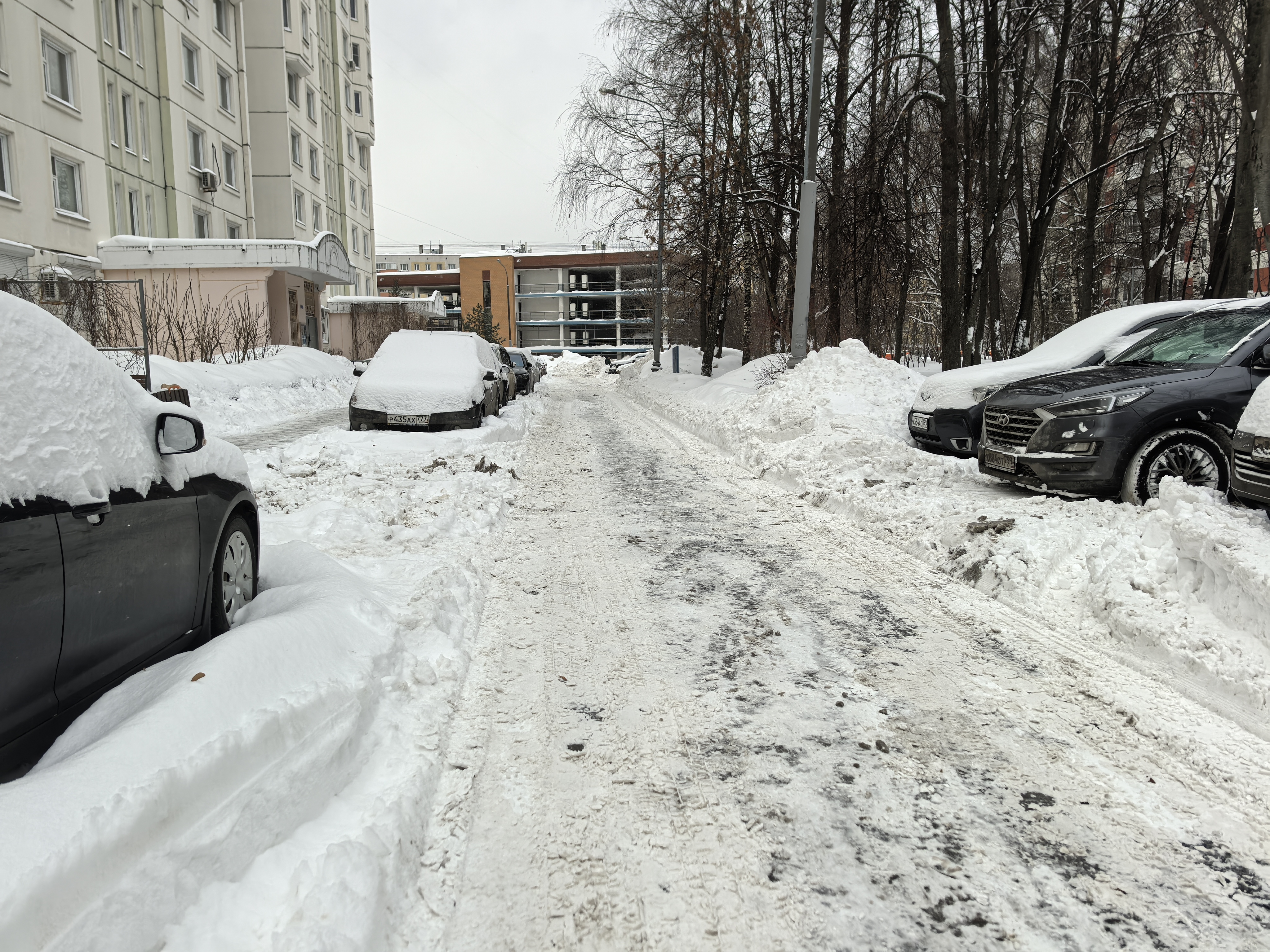 Дворовая территория с припаркованными автомобилями, покрытая снегом. Проезжая часть узкая, тротуары не просматриваются или полностью засыпаны снегом.