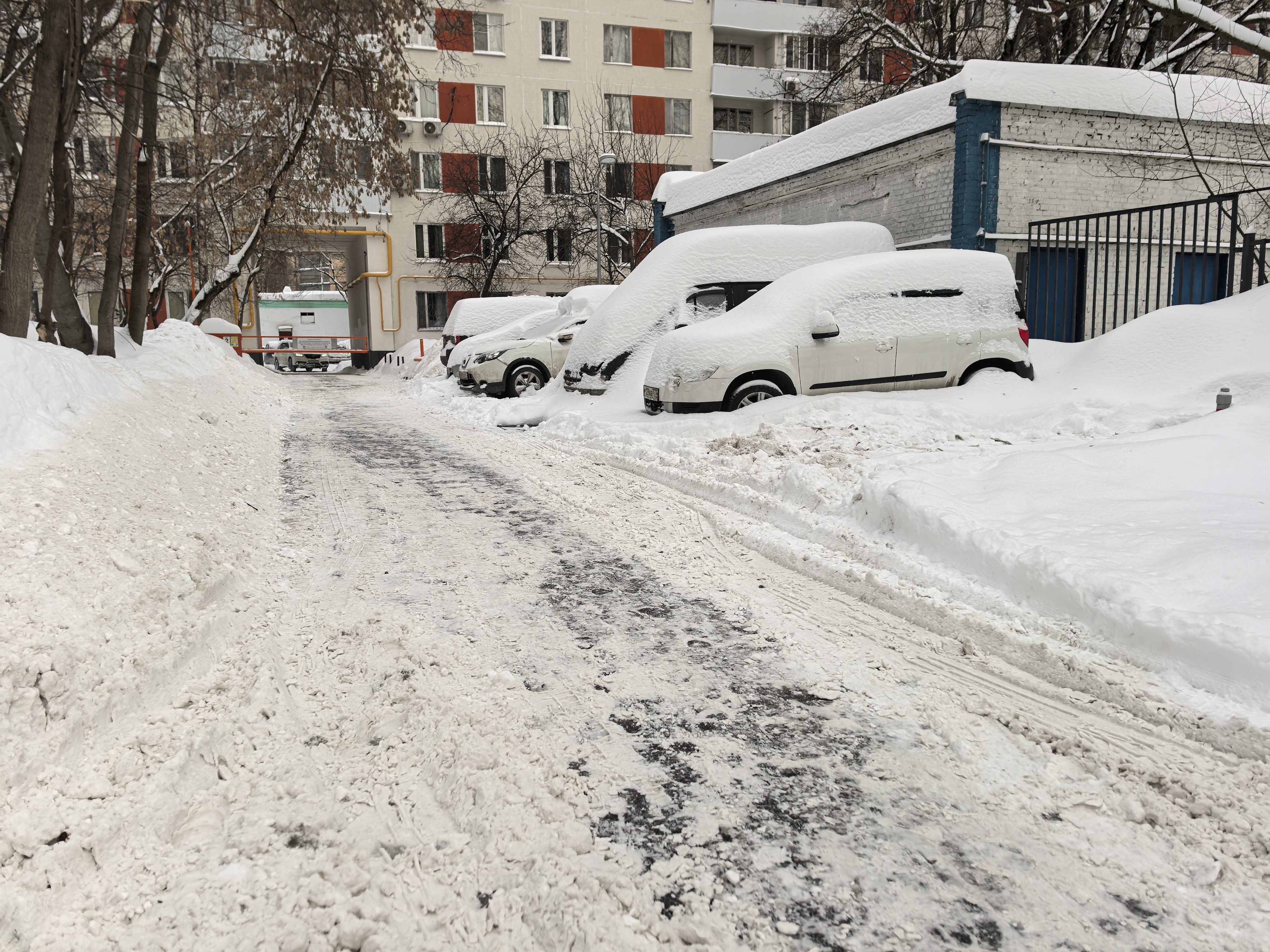 Дворовая территория с припаркованными автомобилями, покрытыми снегом. Проезжая часть частично очищена, но покрыта снегом и грязью. Тротуаров нет, движение пешеходов затруднено.