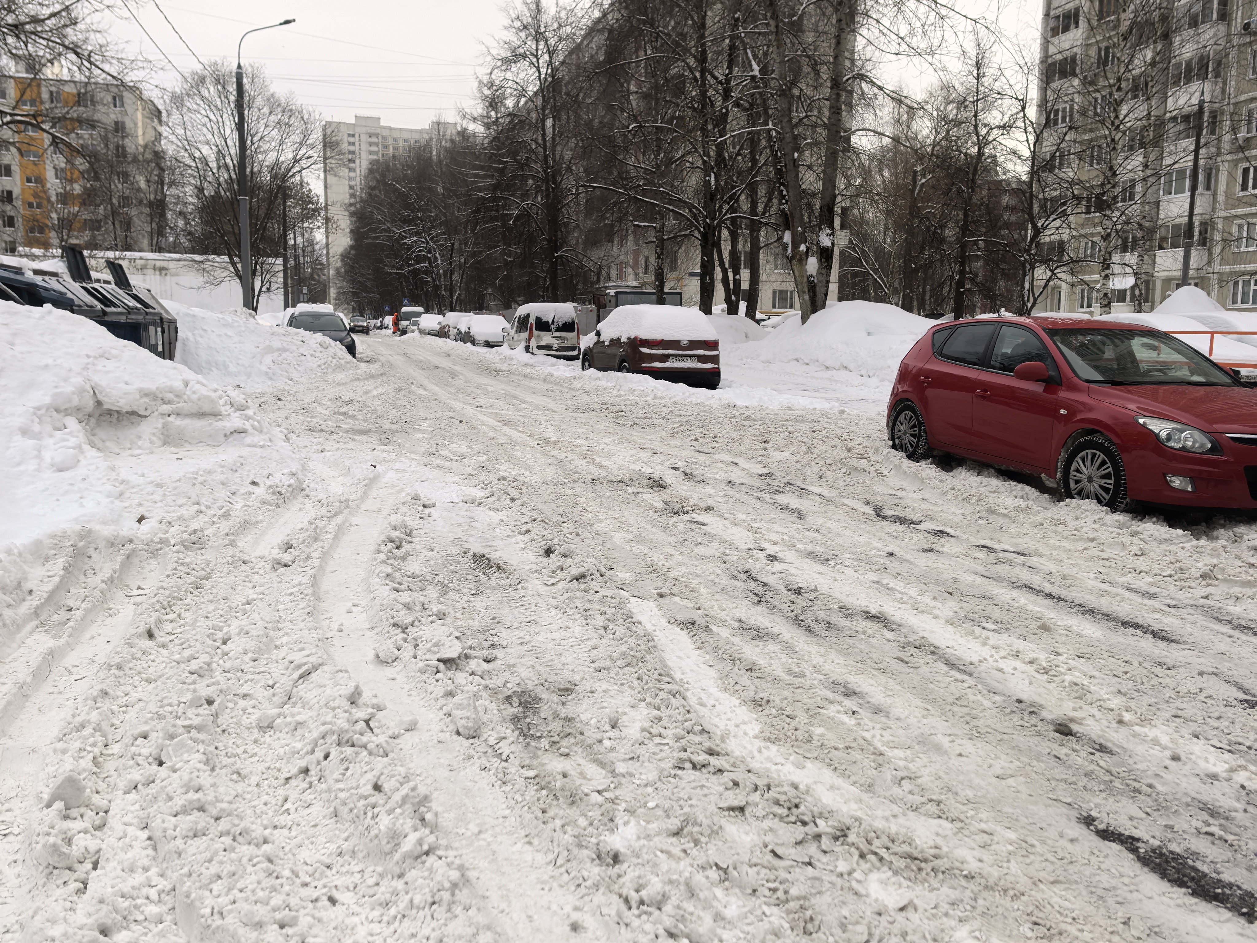 Дворовая территория с припаркованными автомобилями, покрытая снегом и снежной кашей. Видны жилые дома и контейнеры для мусора.