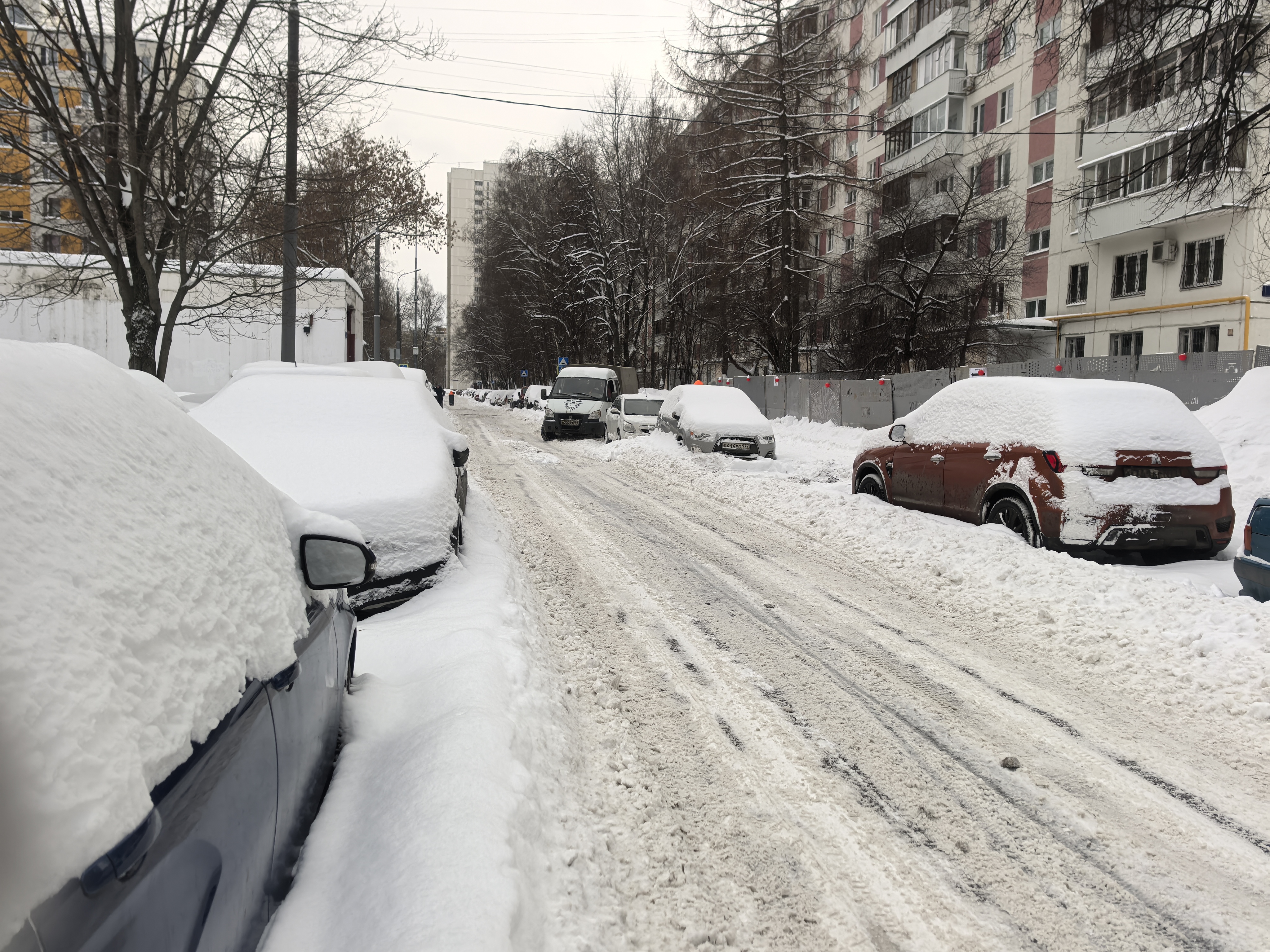 Участок проезжей части в жилой зоне, покрытый снегом и колеей. Пешеходный переход отсутствует. Тротуары полностью заблокированы сугробами, пешеходы вынуждены идти по проезжей части.